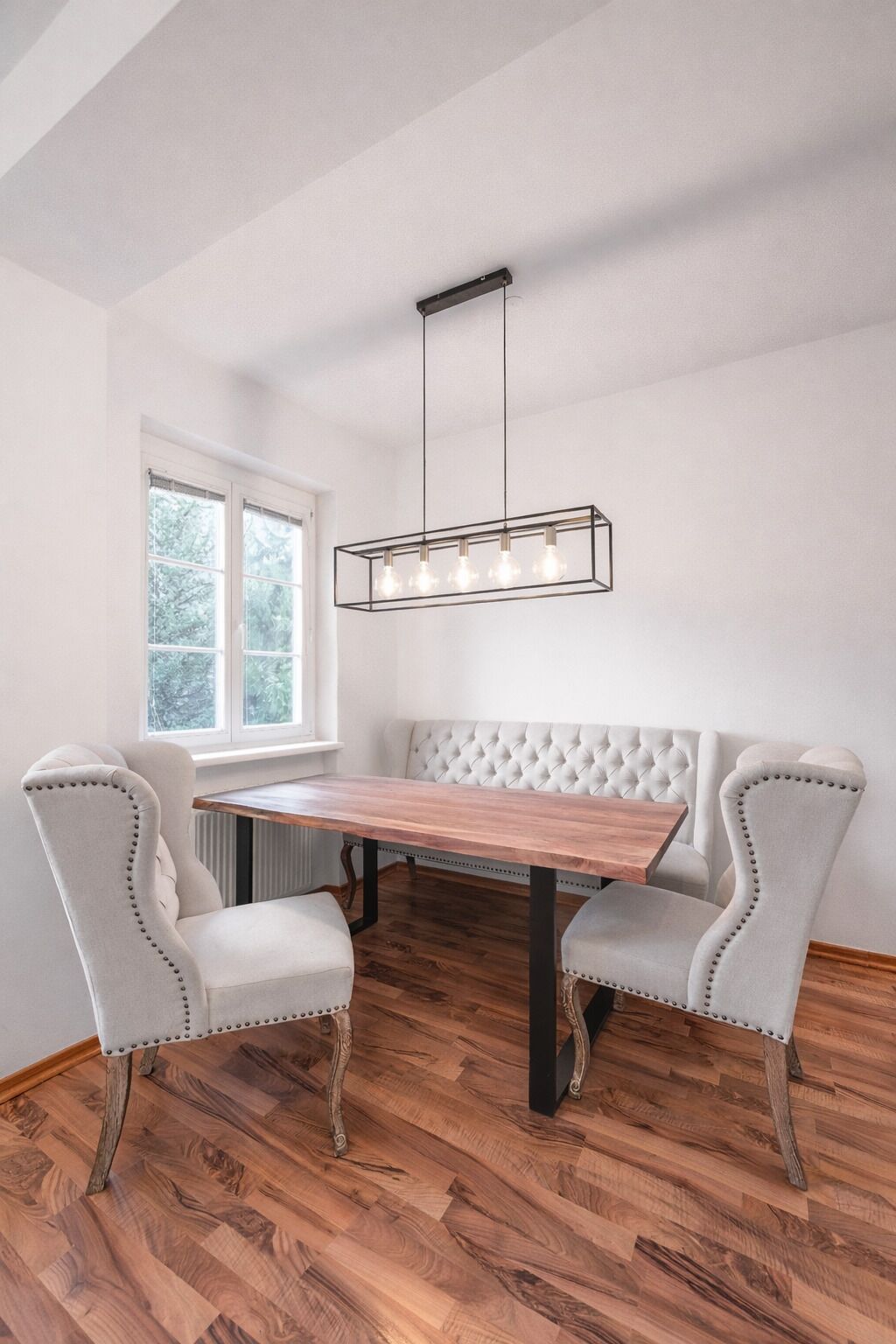 A dining area with a wooden table and two upholstered chairs, a hanging light fixture, and a window with a view of trees.