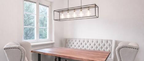 A dining area with a wooden table and two upholstered chairs, a hanging light fixture, and a window with a view of trees.