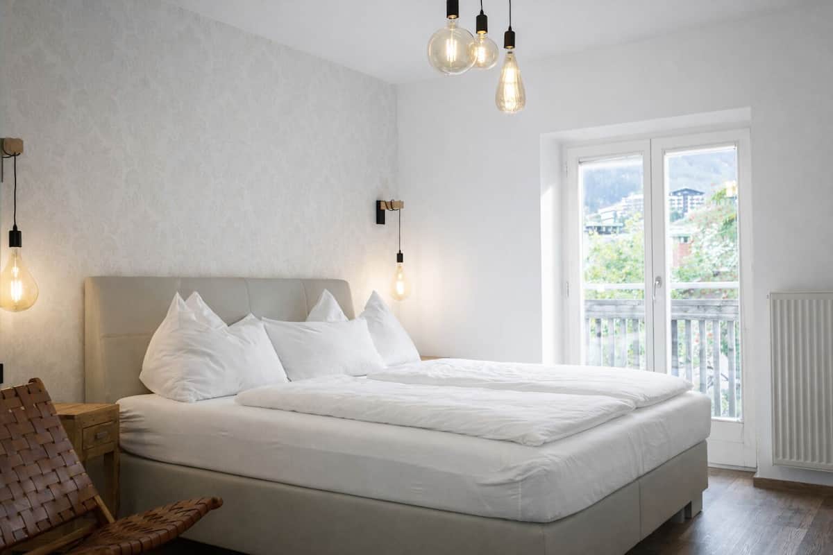 Ein Schlafzimmer mit einem großen Bett, einem Nachttisch aus Holz, einem gewobenen Sessel und Blick ins Freie durch eine Schiebetür.