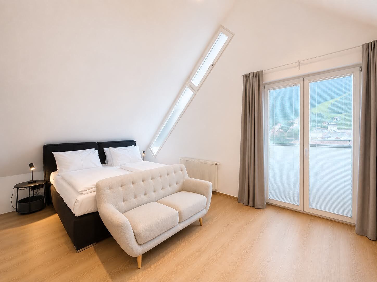 A bedroom with a bed, a sofa, a nightstand, a lamp, a window with curtains, and a view of the outside.