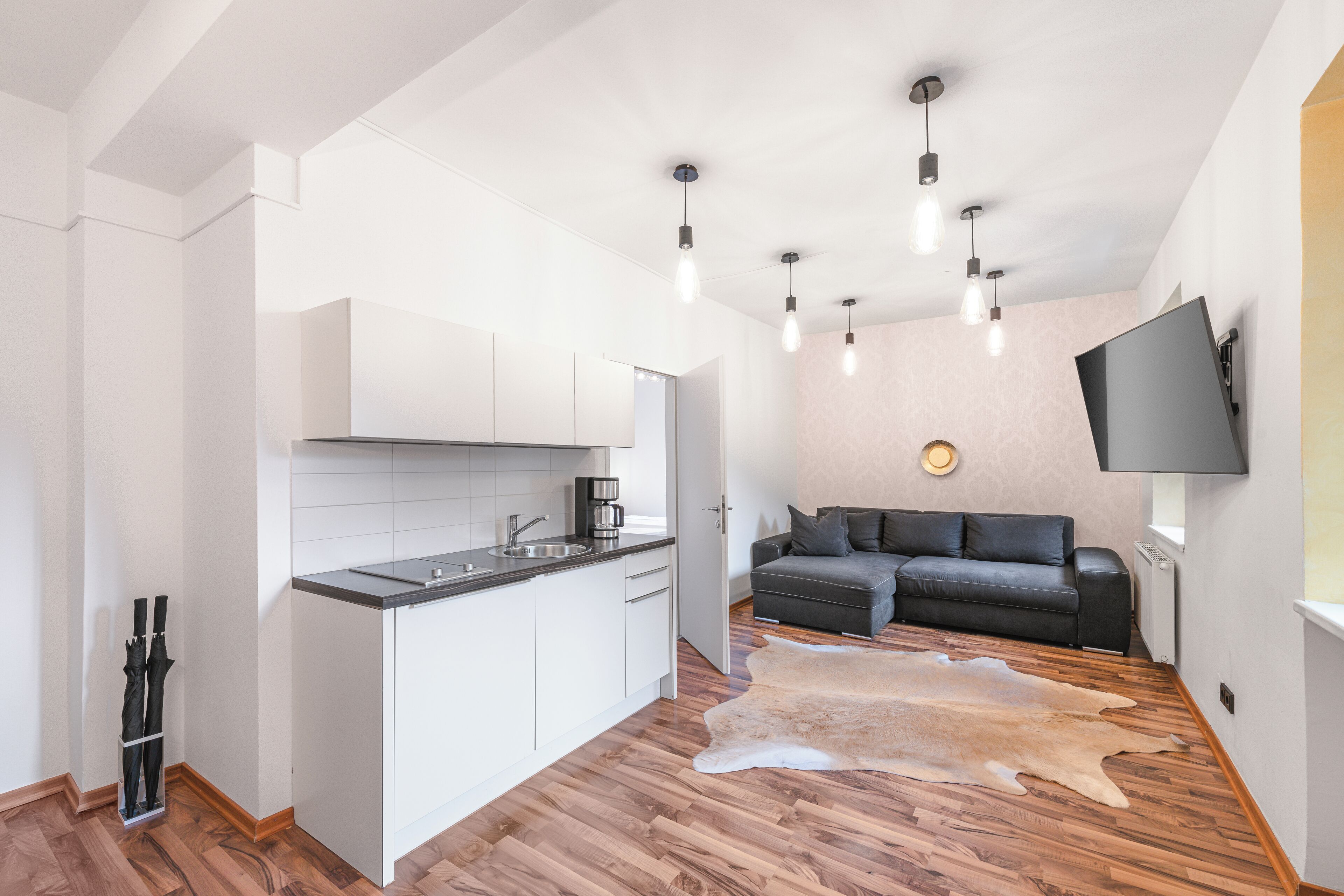 A modern kitchen with white cabinets and a wooden floor, a grey sofa, and a mounted TV in a living area.