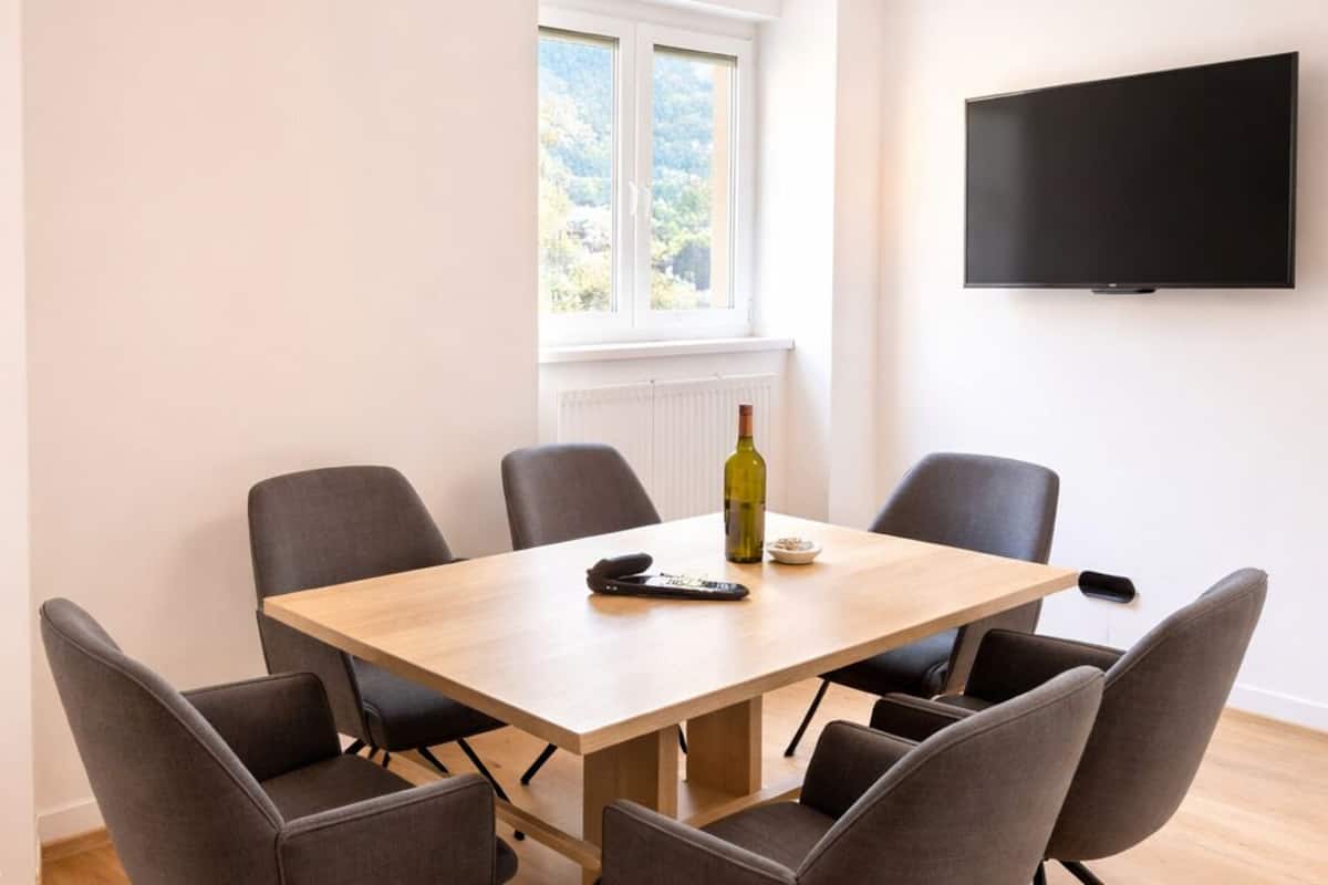 Ein Konferenzraum mit einem Holztisch, sechs grauen Stühlen, einer Weinflasche, einer Schüssel und einer Fernbedienung.
