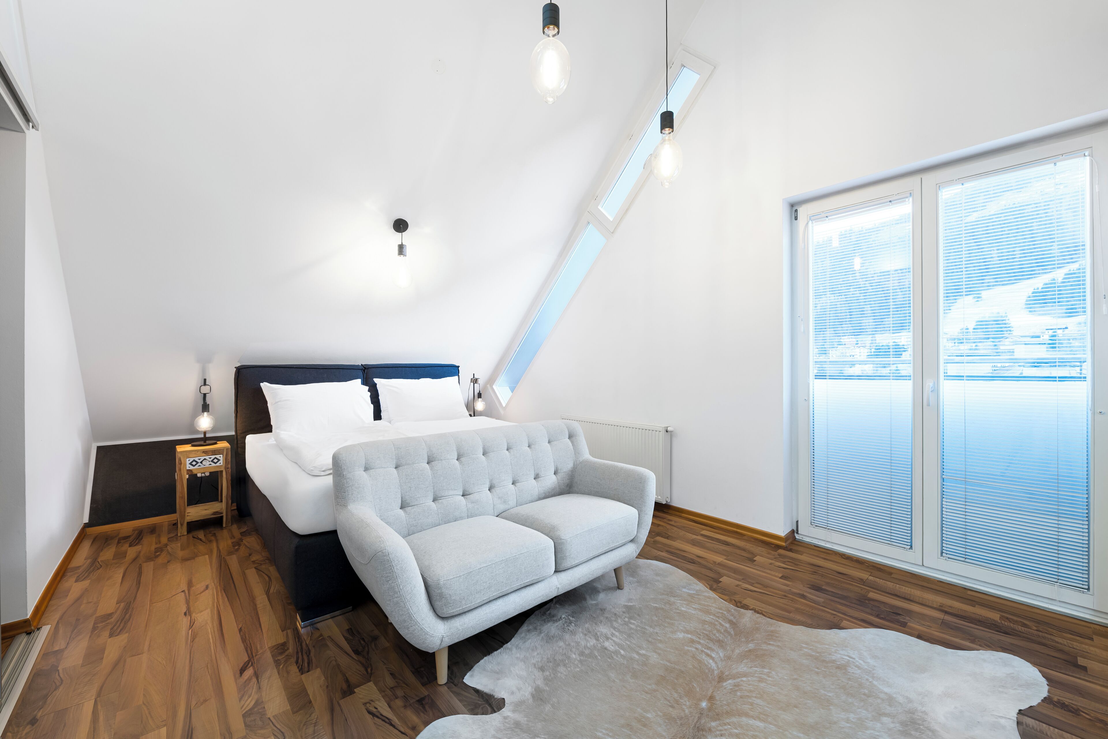 A bedroom with a bed, a grey sofa, a small wooden side table, and a large window with blinds.