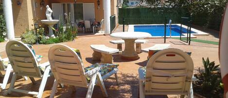 An outdoor patio with white plastic chairs and a table, a small fountain, and a swimming pool.
