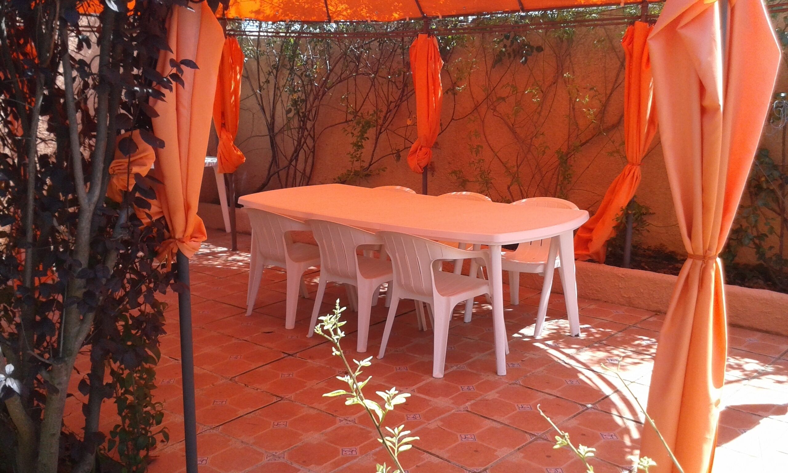 An outdoor patio with a white table and chairs, surrounded by orange curtains and greenery.