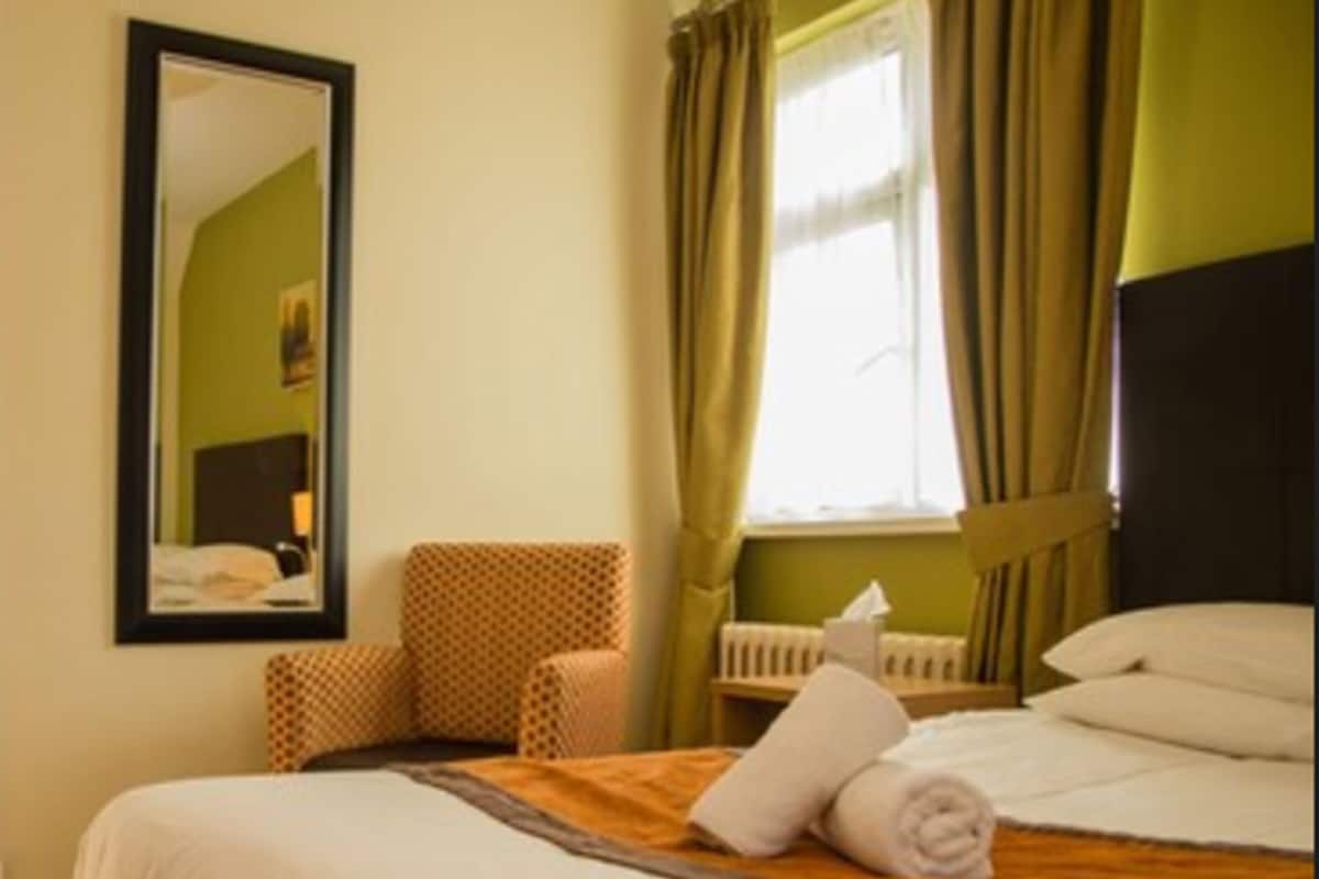 Una habitación de hotel con cama, una silla, una ventana con cortinas y un espejo en la pared.