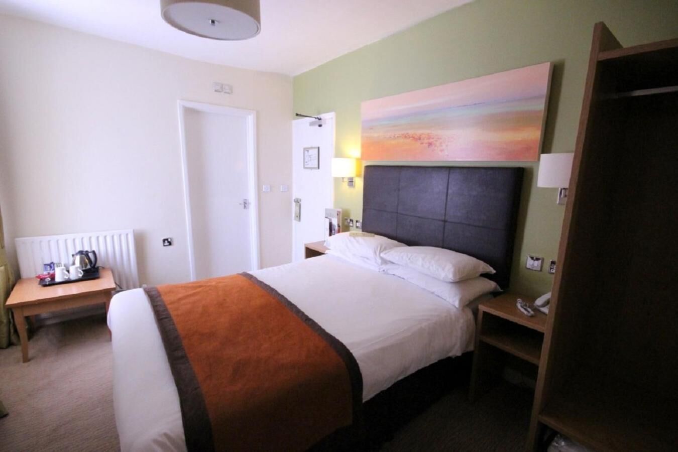 Habitación de hotel con cama, mesitas de noche, escritorio con tetera y un cuadro en la pared.