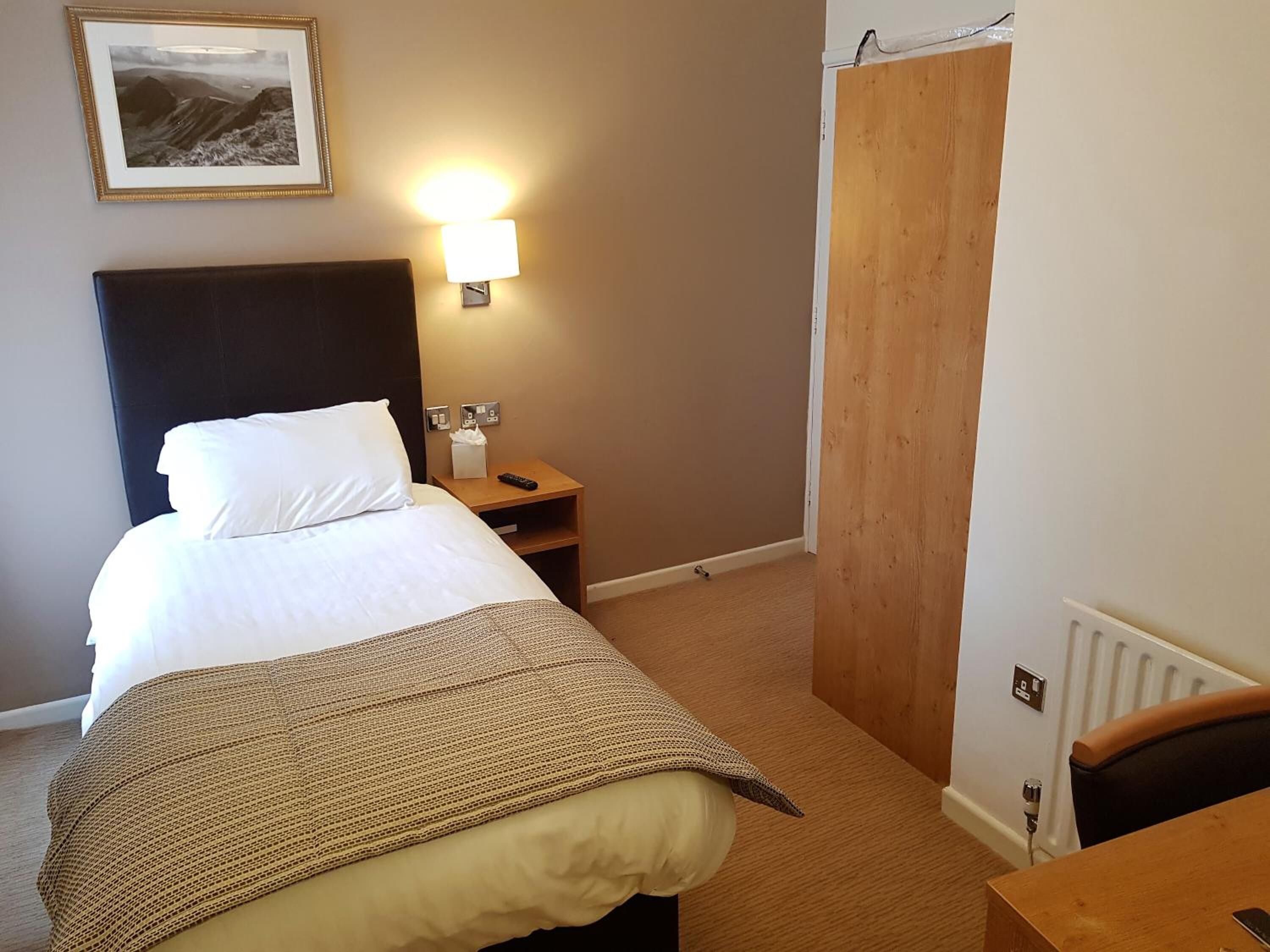 A hotel room with a bed, a bedside table with a lamp, a wooden wardrobe, and a framed picture on the wall.