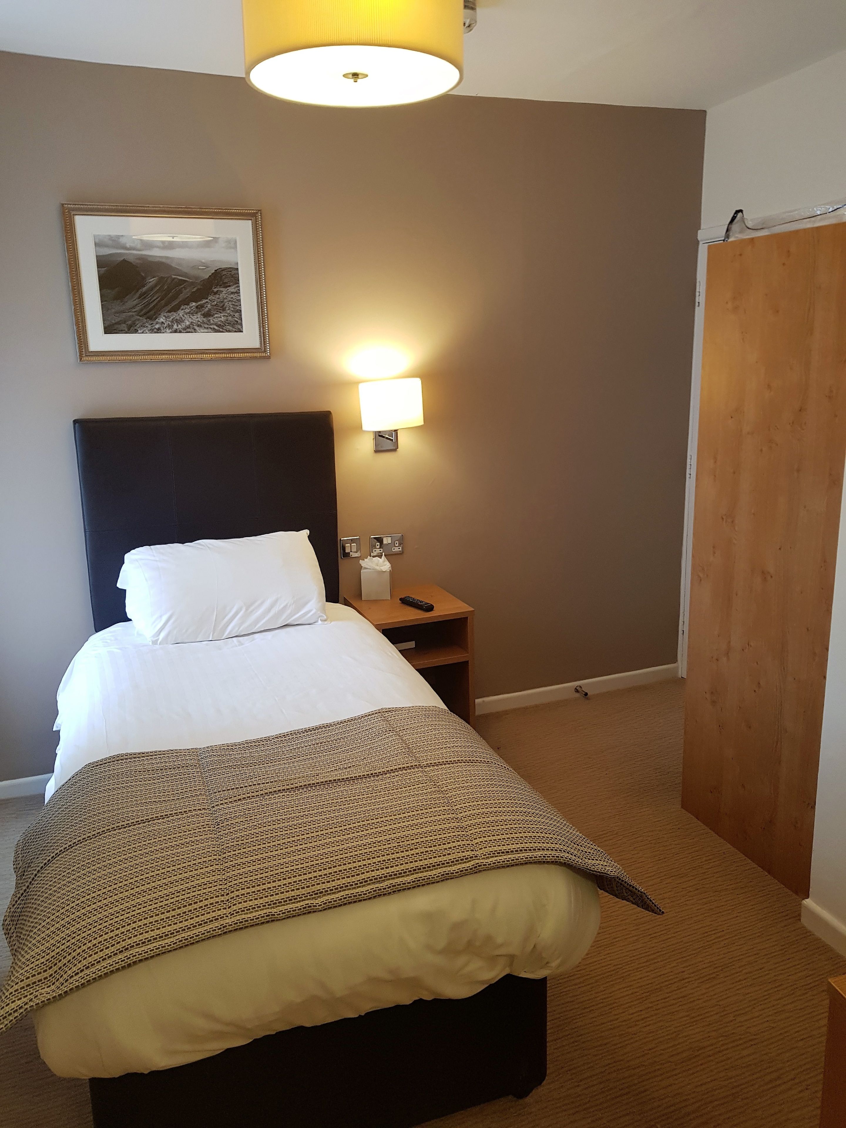 A neatly made bed with a white comforter and a brown knitted throw, a bedside table with a remote control, a wall-mounted lamp, and a framed picture on the wall.