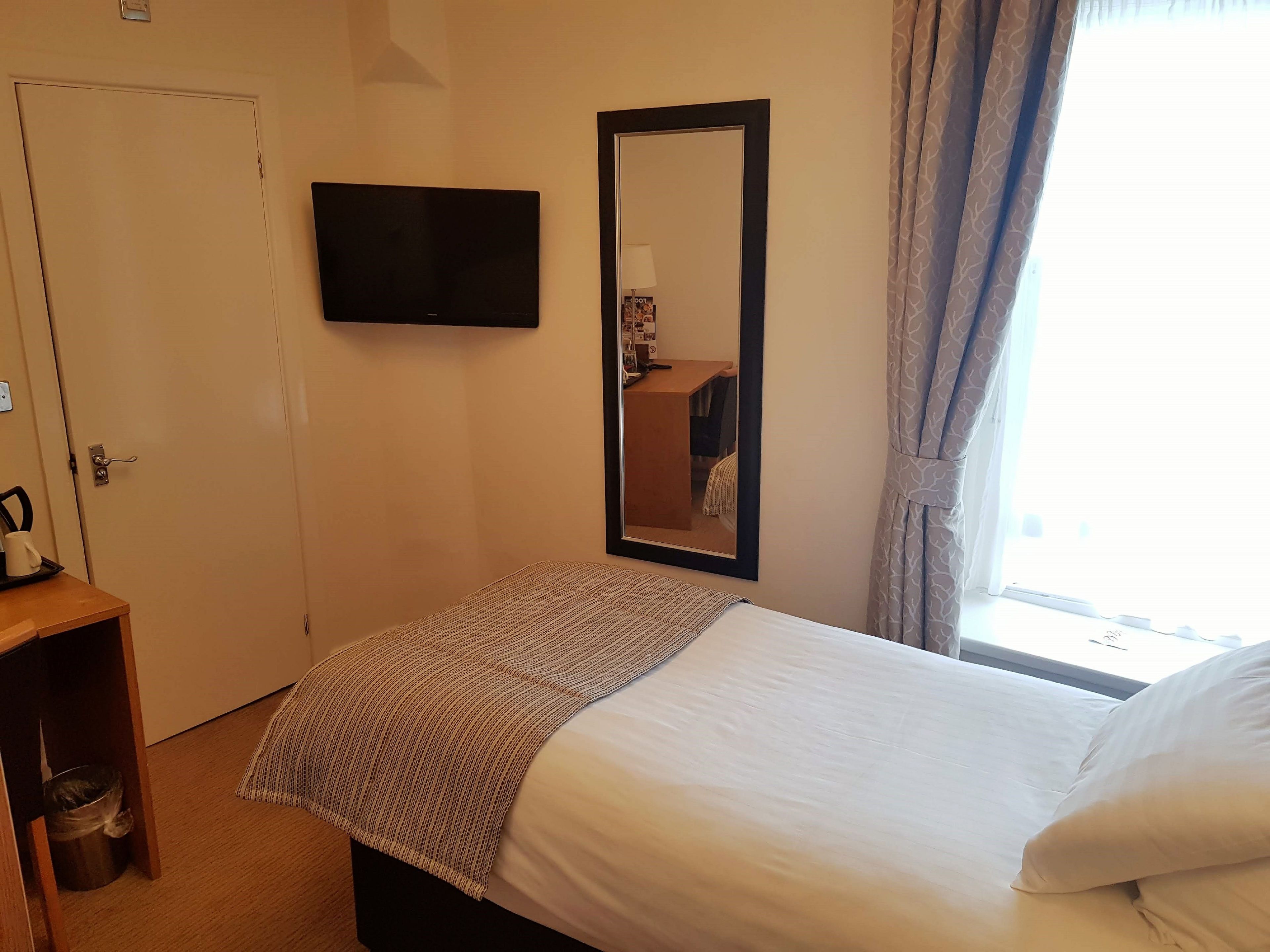 Habitación de hotel con cama, televisor montado en la pared, una mesita con hervidor y ventana con cortinas.