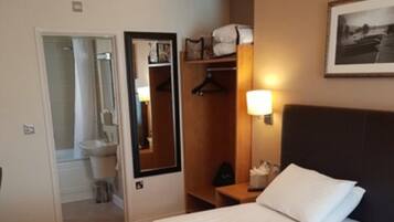 Quarto de hotel com uma cama, mesa de cabeceira, espelho, guarda-roupa e banheiro visível através de uma porta aberta.