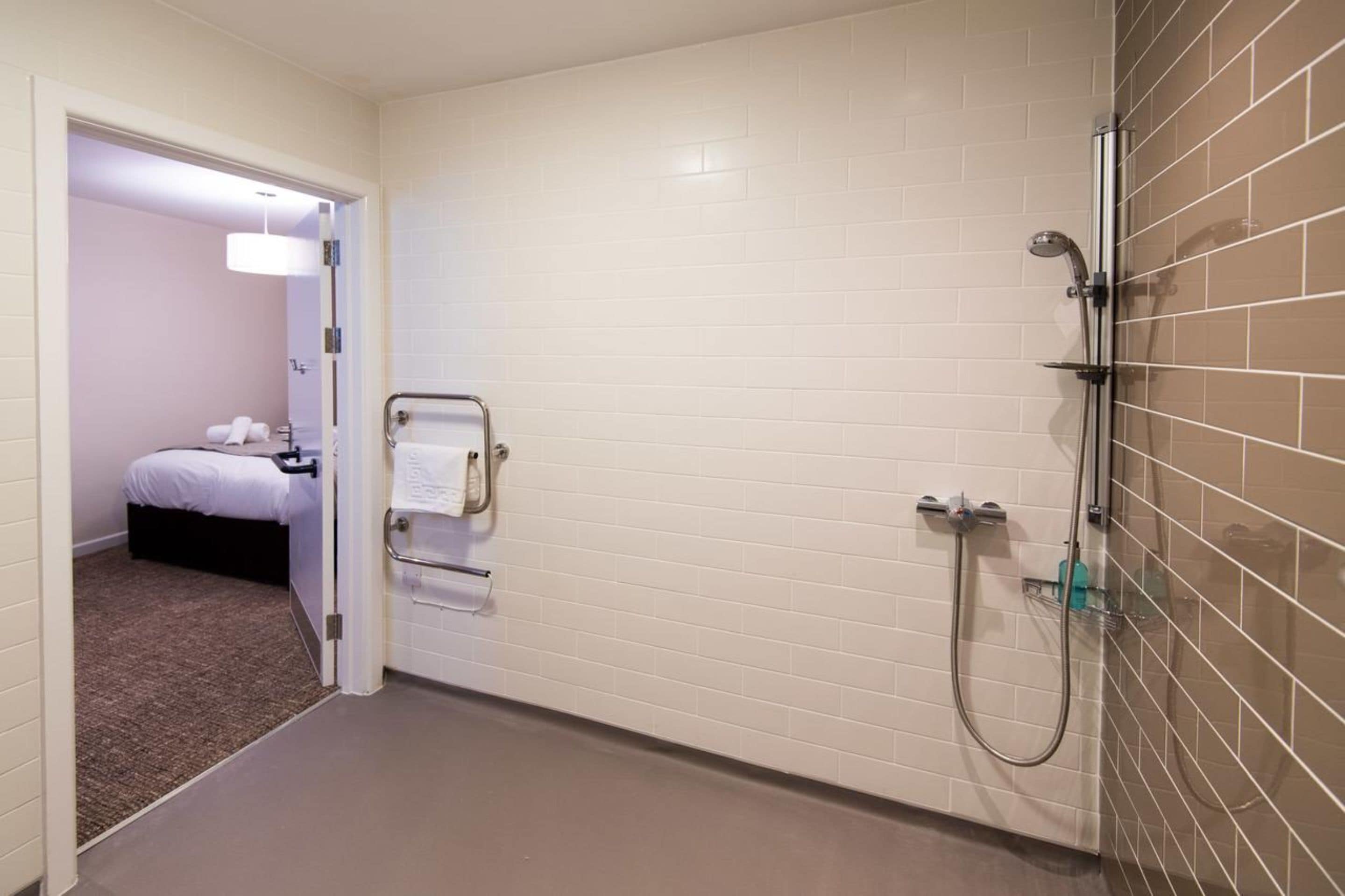 Een hotelbadkamer met witte betegelde wanden, een douche, een handdoekhouder en een zichtbare deur naar een slaapkamer.