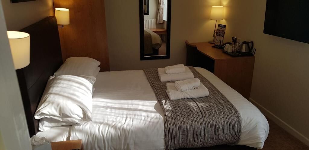 Una habitación de hotel con una cama bien hecha, dos lámparas de noche, una mesita de noche con lámpara, un espejo y un televisor de pantalla plana.