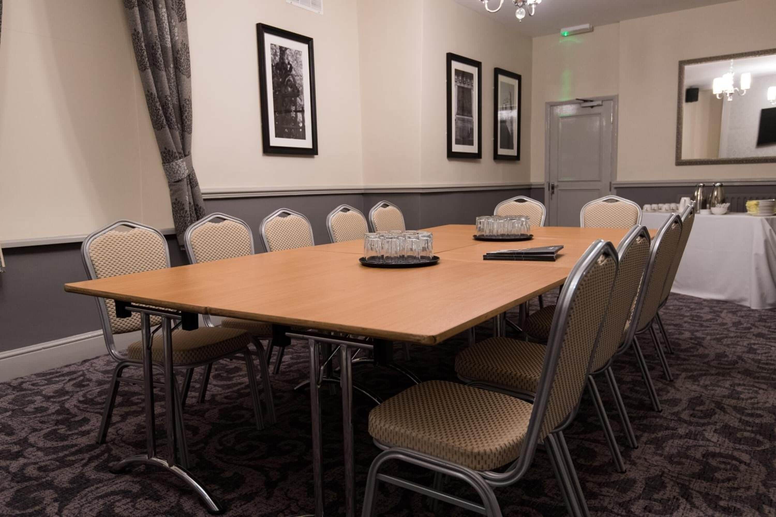 A conference room with a long table, chairs, framed pictures on the wall, and a door in the background.