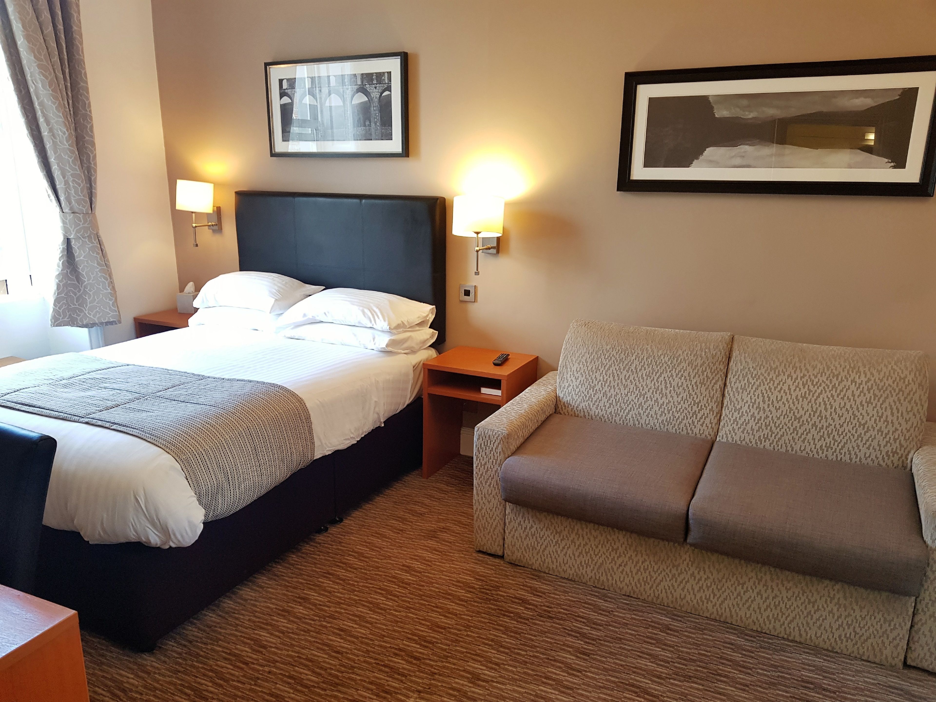 A hotel room with a bed, a sofa, two wall-mounted lamps, and two framed pictures on the wall.