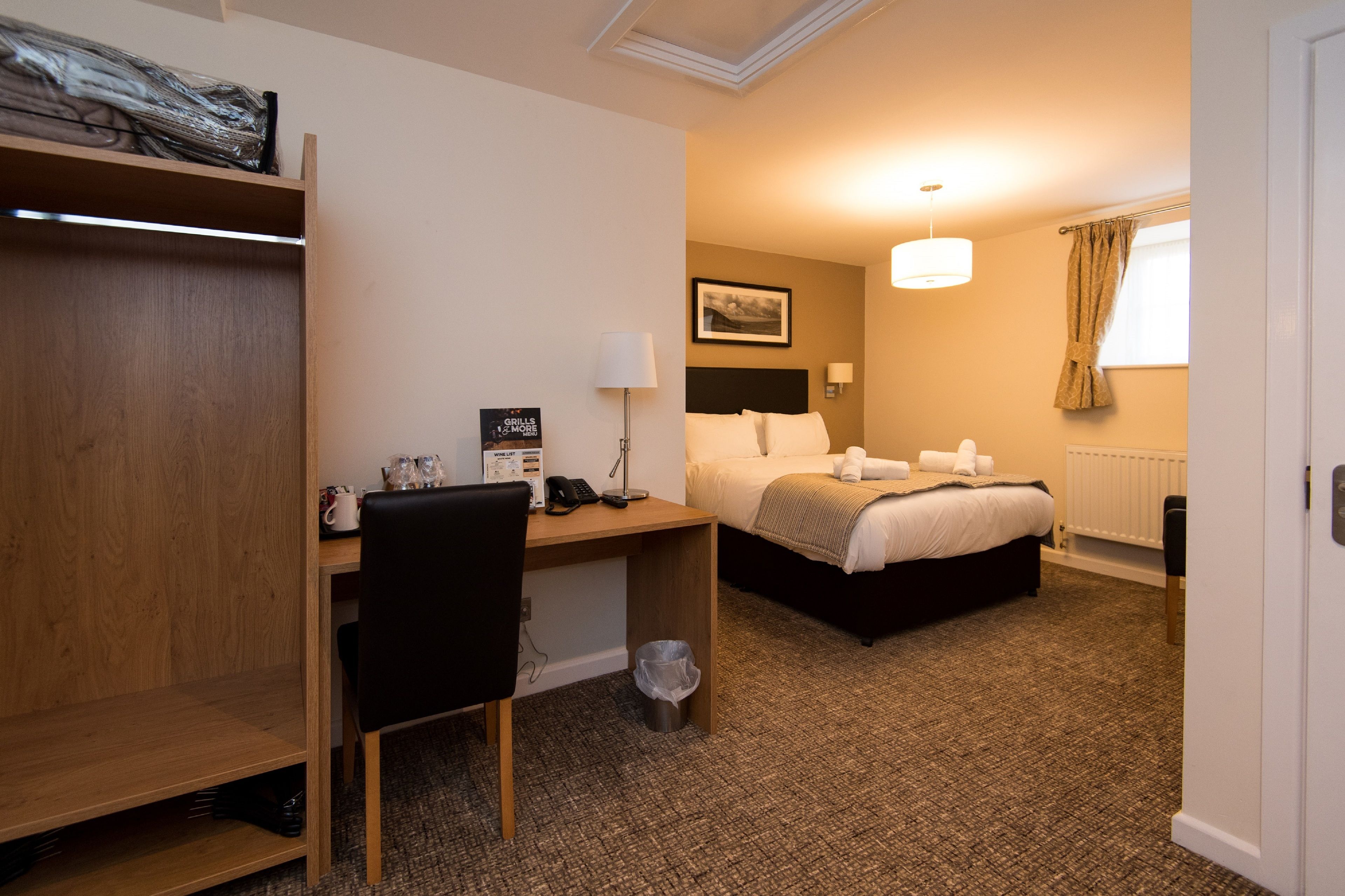 A hotel room with a wooden desk, a bed, a chair, a lamp, and a picture on the wall.