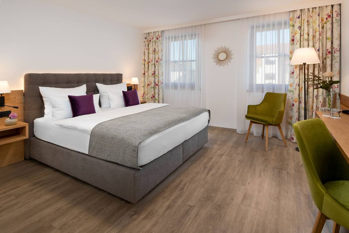 A hotel room with a large bed, a green chair, a wooden nightstand, and a desk with a lamp.