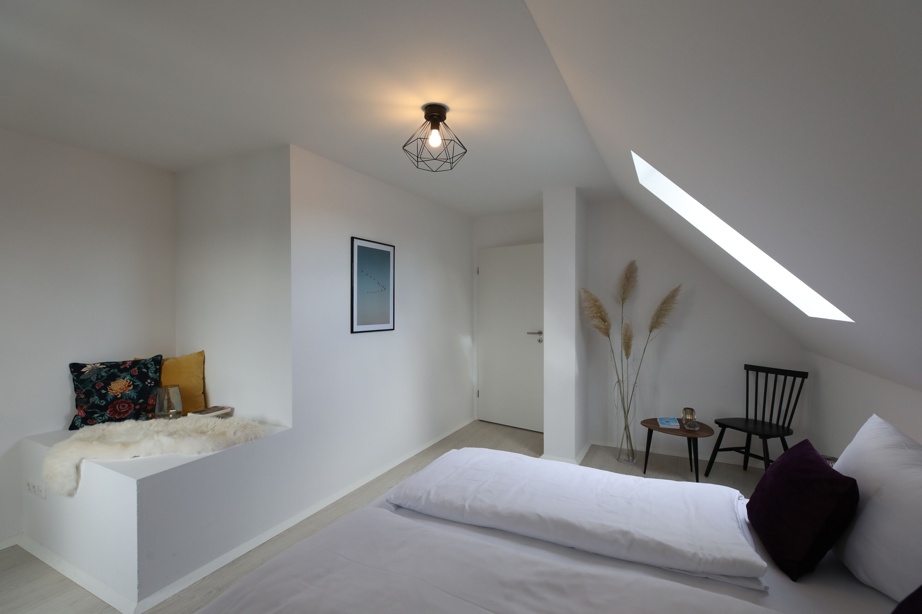 A modern bedroom with a loft-style ceiling, a bed, a small seating area, and a skylight.