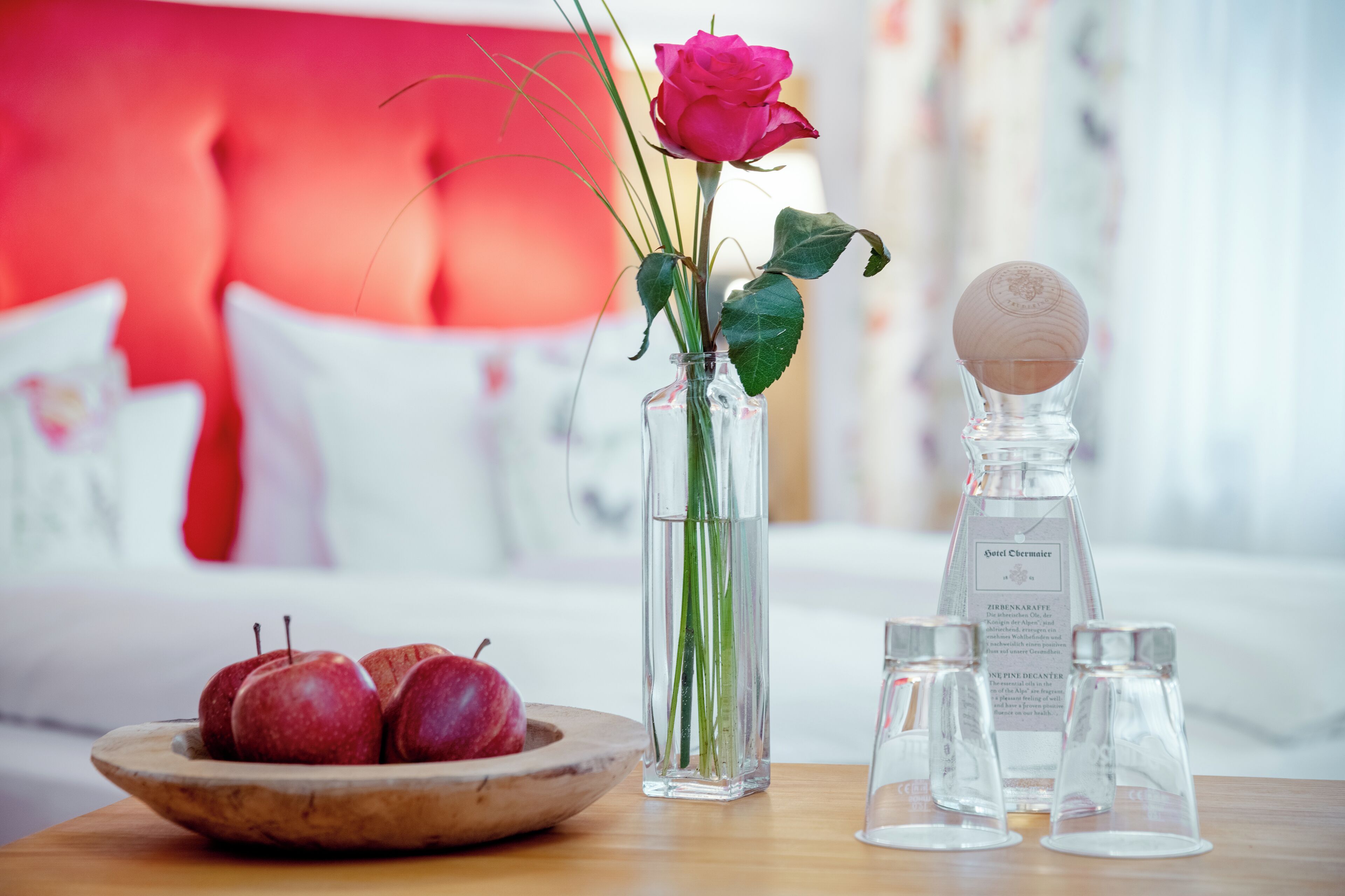 Una mesa con un cuenco de madera lleno de manzanas, un florero con una rosa y tallos verdes, y tres botellas de vidrio transparente con etiquetas.