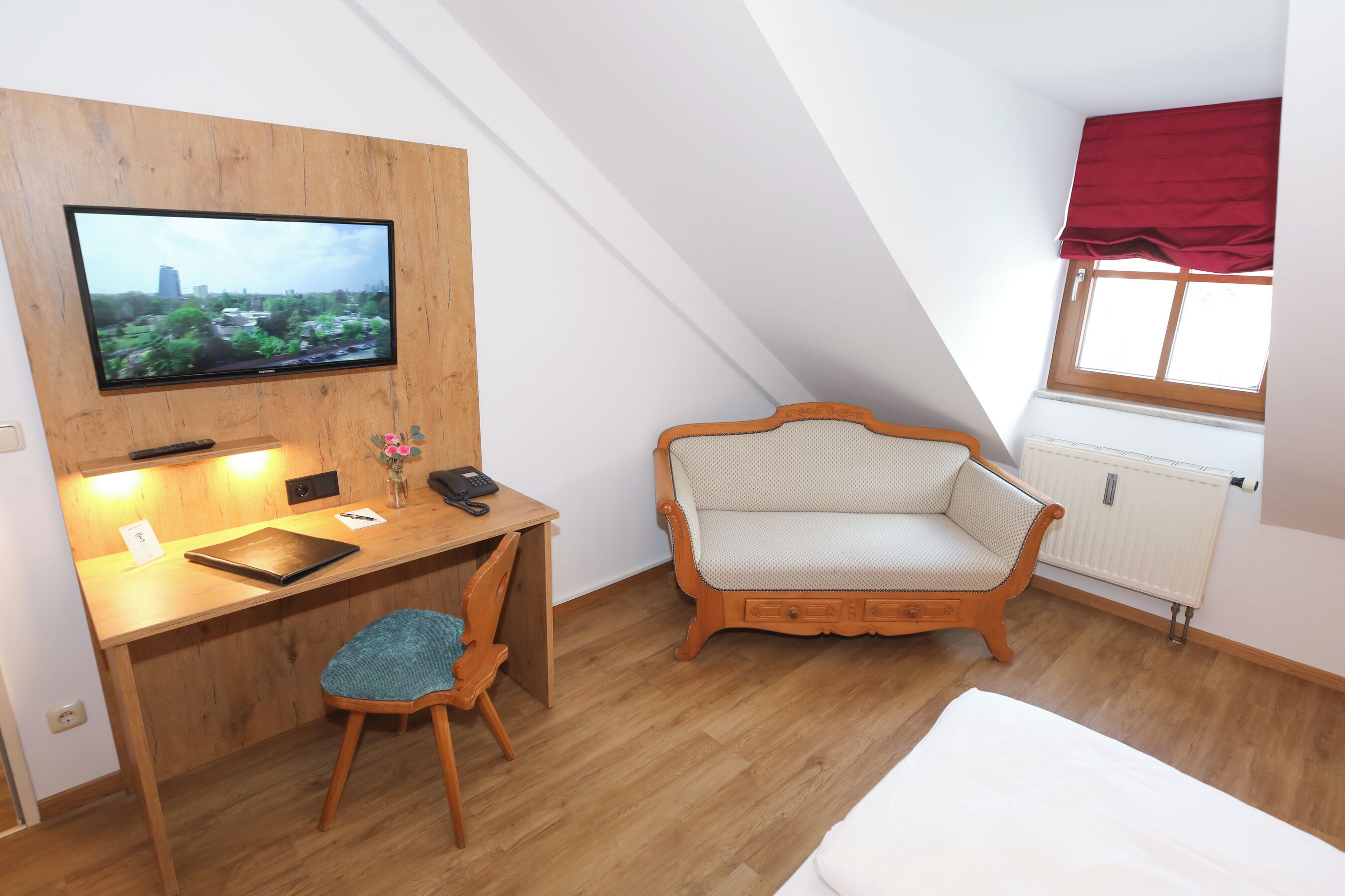 A room with a wooden desk, a chair, a bed, a TV mounted on the wall, and a window with a red valance.