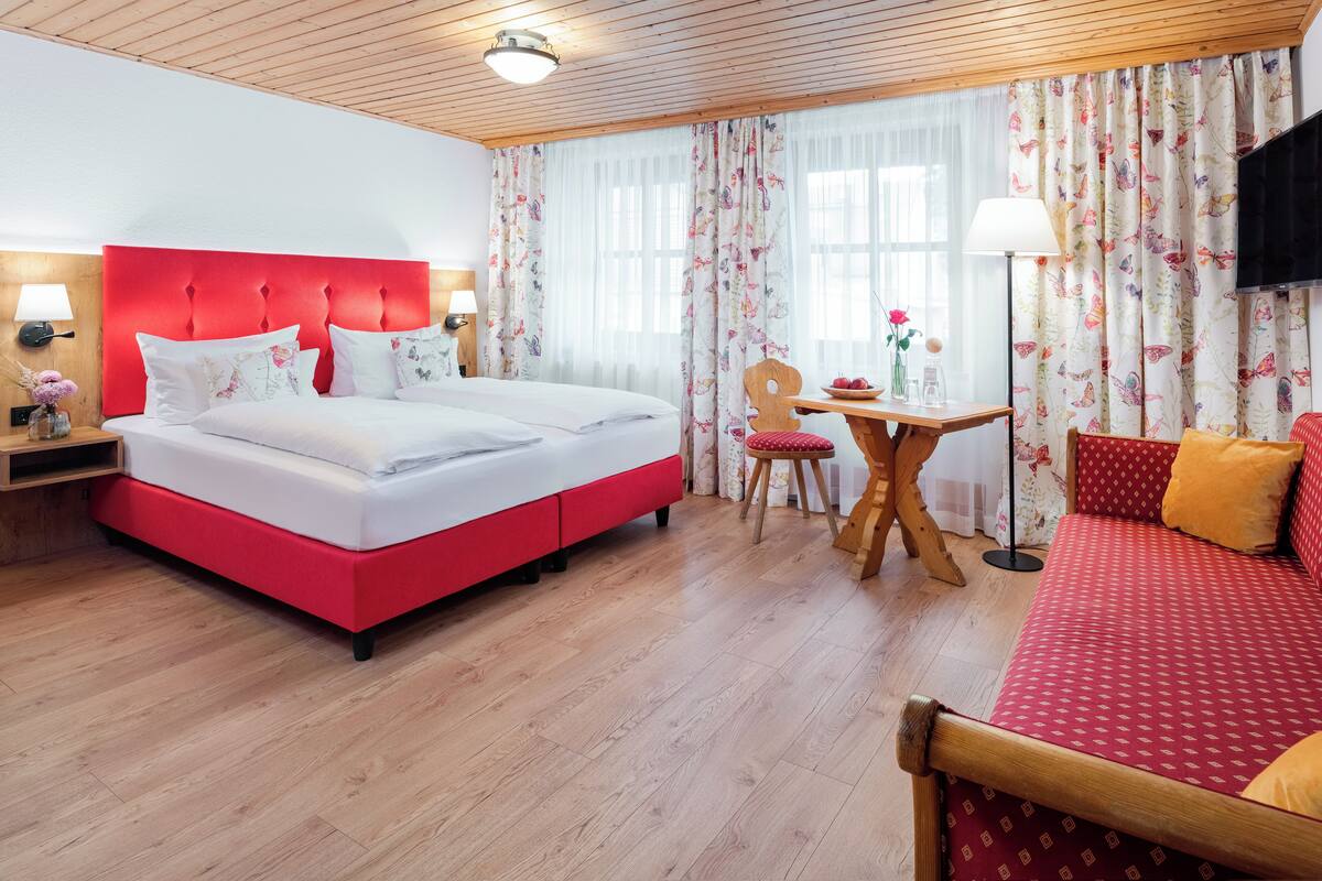A hotel room with a red bed, a wooden headboard, a wooden floor, a small table with a chair, a sofa, and a TV on the wall.