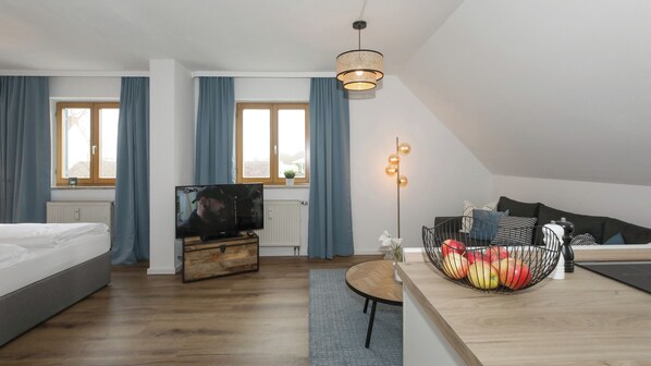 A modern hotel room with a bed, TV, wooden dresser, and a dining area with a fruit bowl. - Hotel Obermaier (Munich)