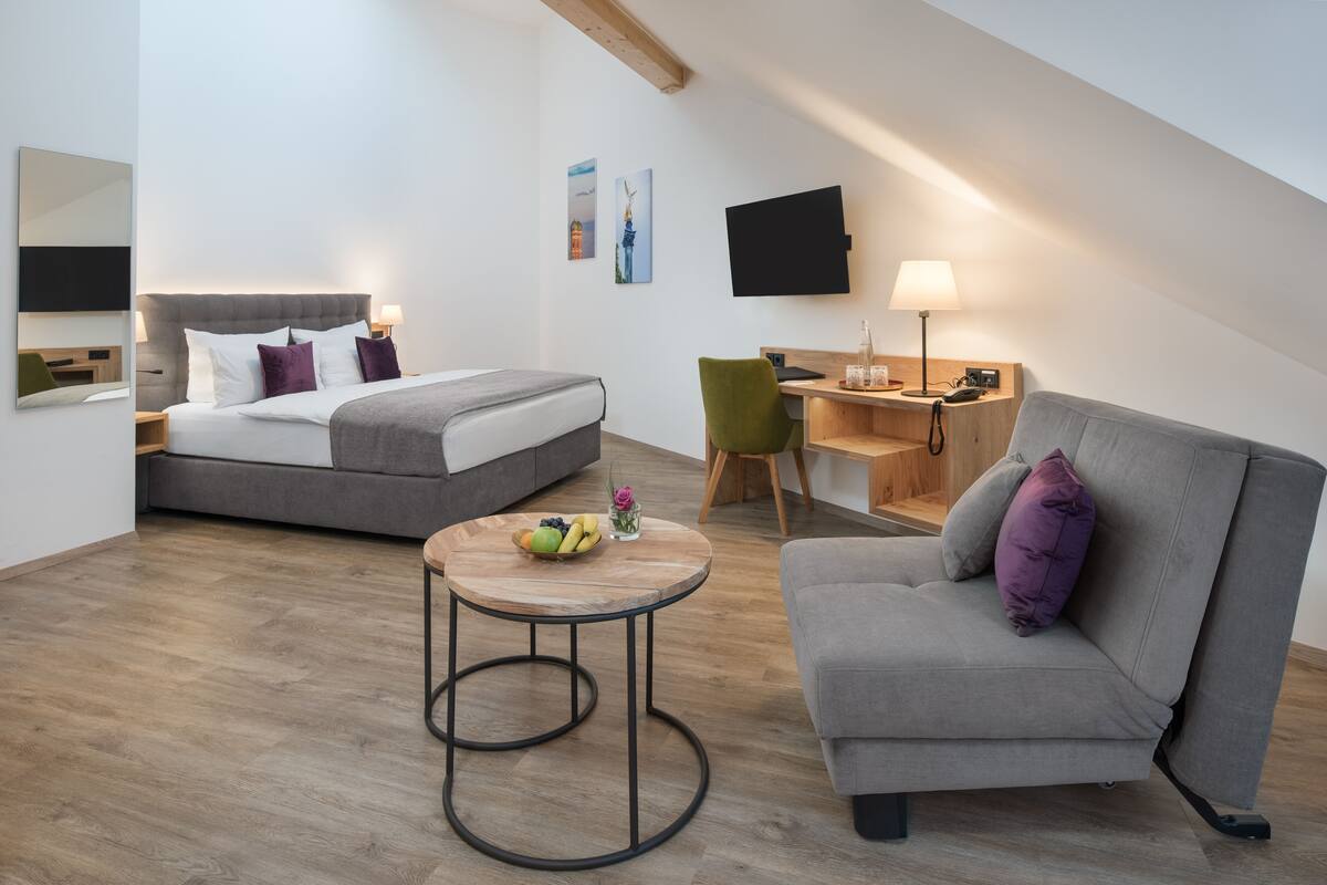 A modern, well-lit room with a bed, a sofa, a desk, and a round table with fruit.