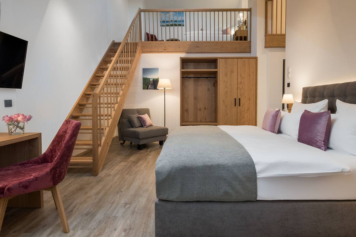 A bedroom with a large bed, a wooden staircase, a desk, and a chair.