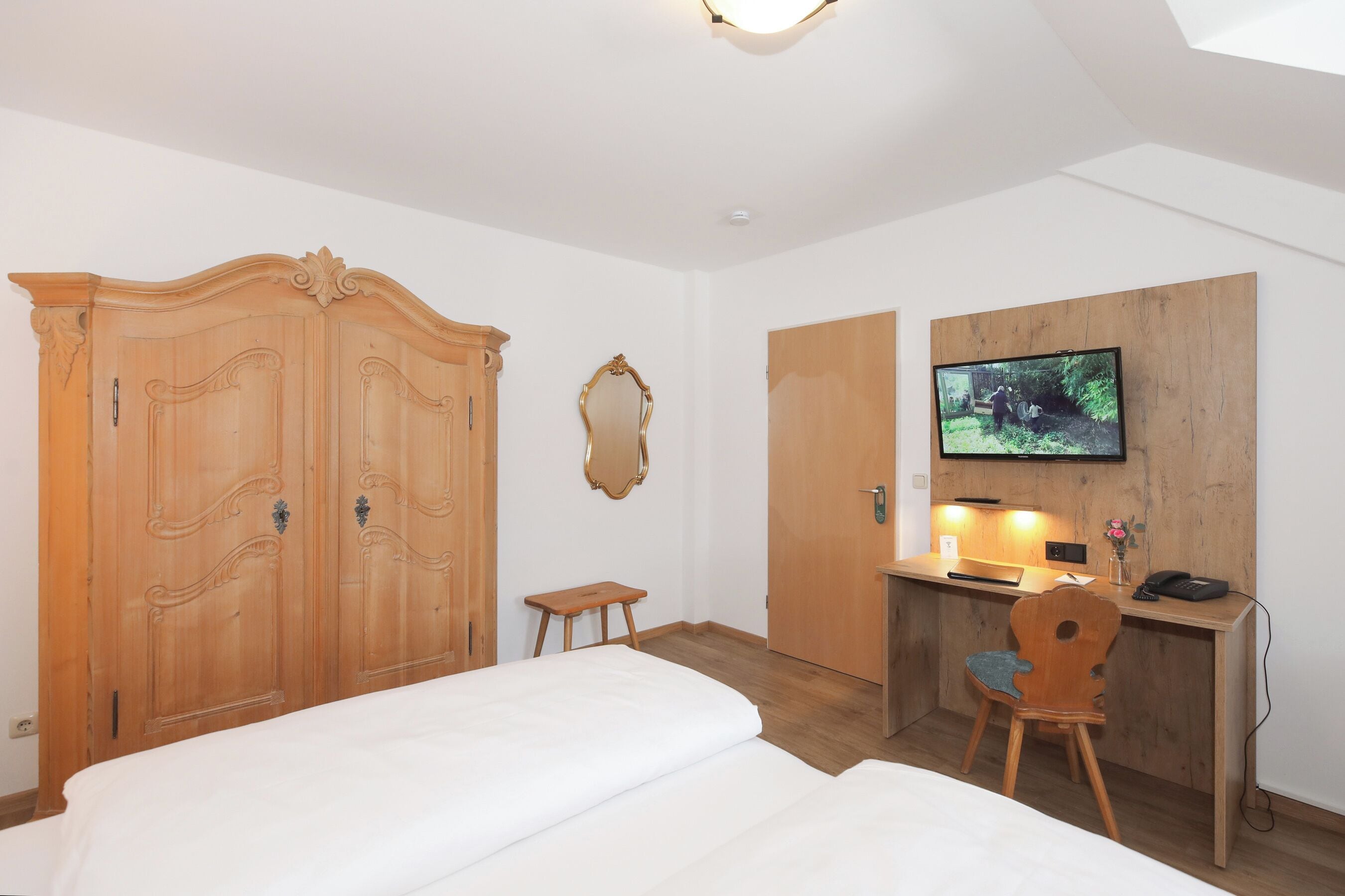 A bedroom with a bed, a wooden wardrobe, a desk with a chair, a television, and a small stool.
