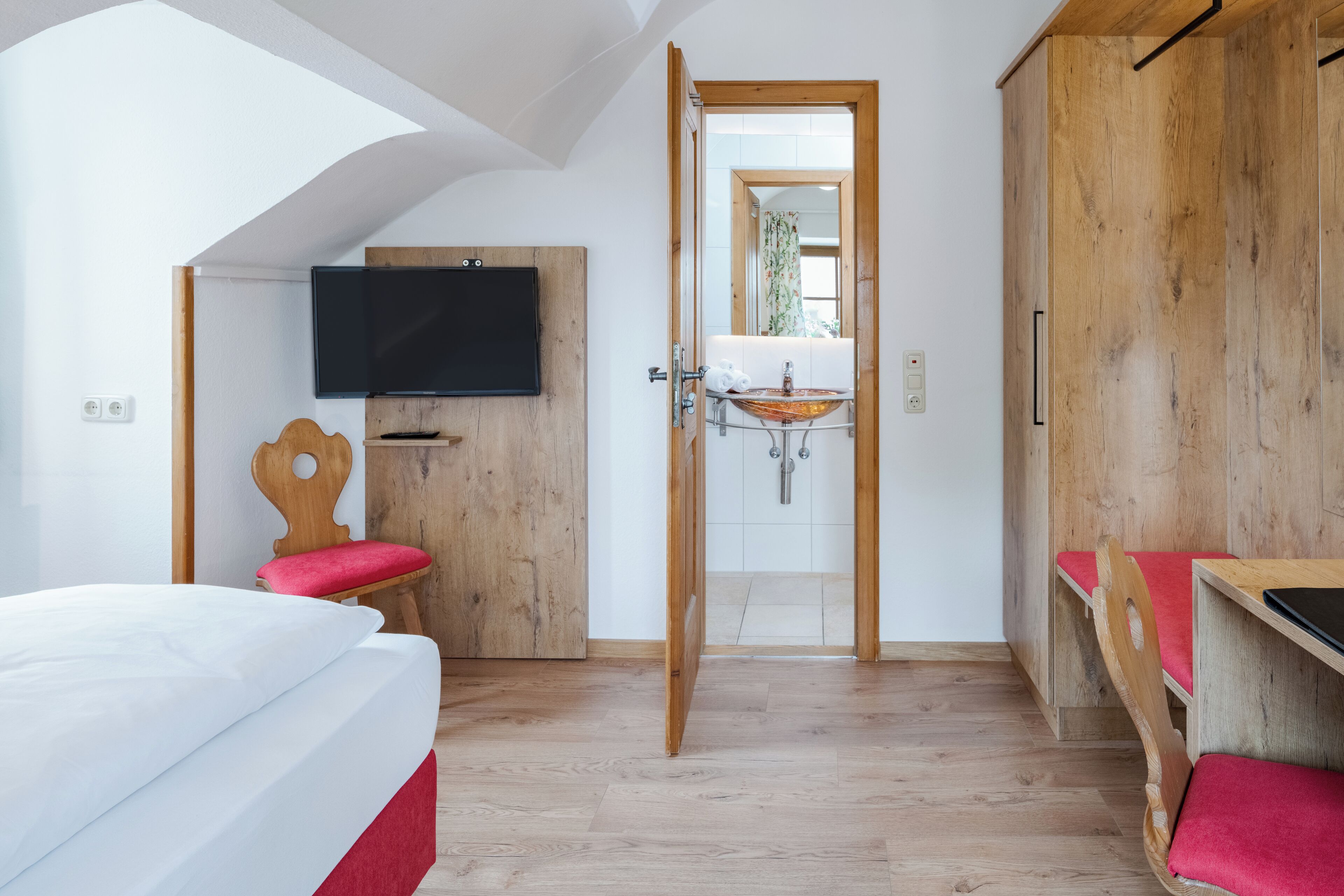 A bedroom with a bed, a TV mounted on a wooden wall, a wooden wardrobe, and a bathroom visible through an open door.