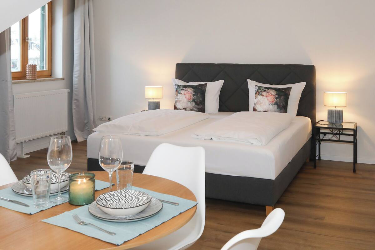 A bedroom with a bed, two bedside lamps, a nightstand, a table with a candle, glasses, and a bowl.