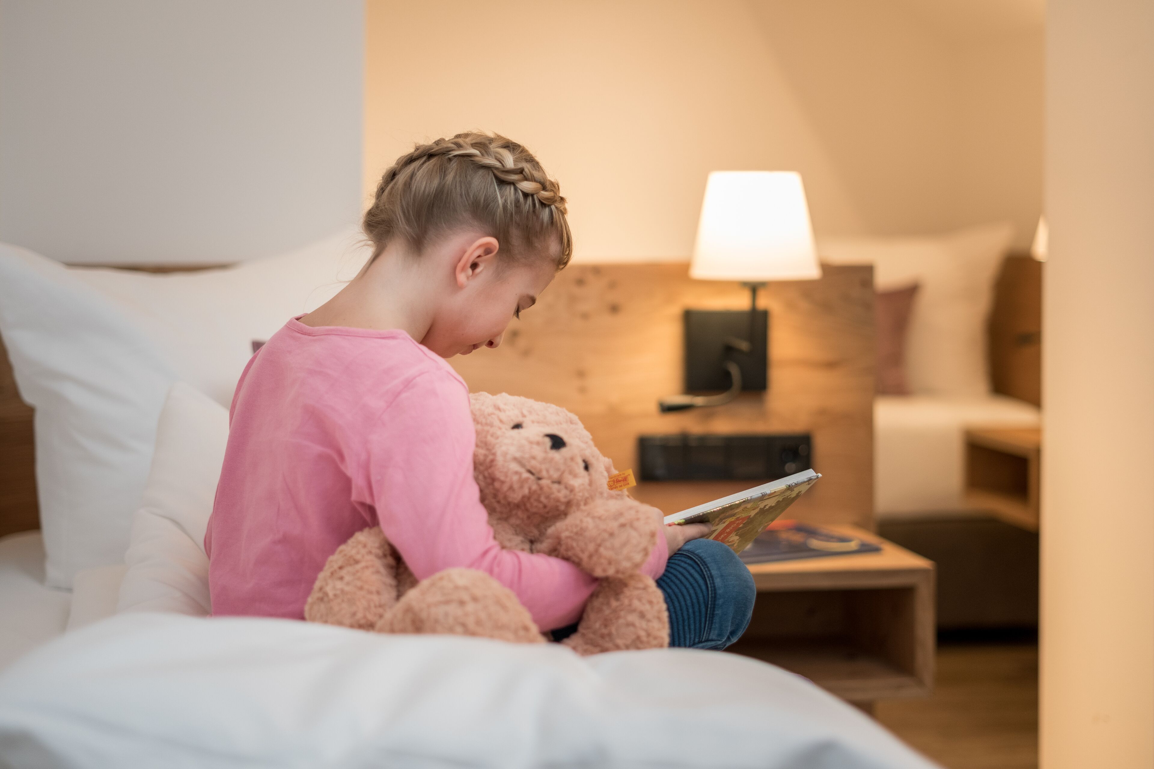 Un niño sentado en una cama con un osito de peluche, leyendo un libro.