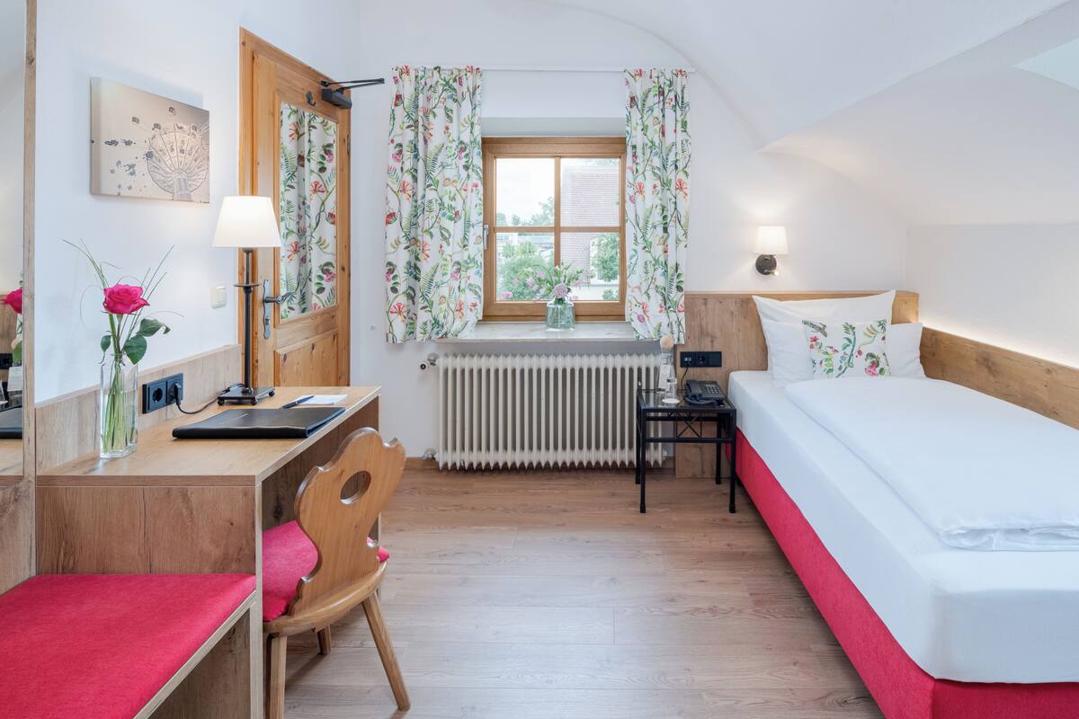 A bedroom with a wooden desk, a chair, a bed, a window with floral curtains, a radiator, and a bedside table with a lamp.