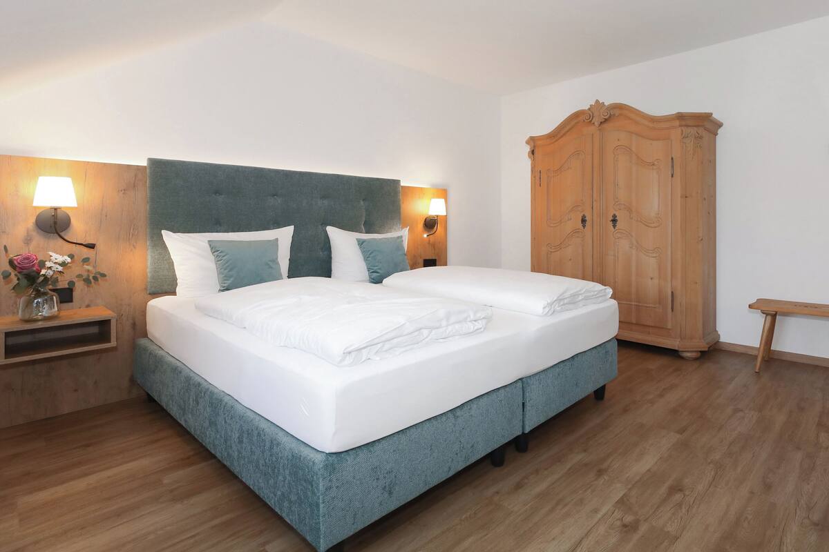 A neatly made bed with white linens and teal bedding, a wooden wardrobe, and a nightstand with a lamp and a vase of flowers.