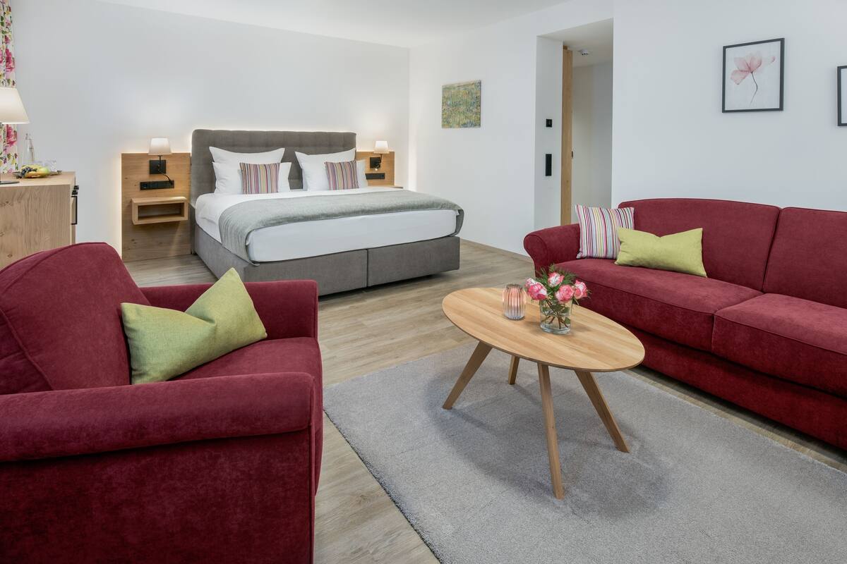 A modern hotel room with a large bed, two red sofas, a wooden coffee table, and a small side table with a lamp.
