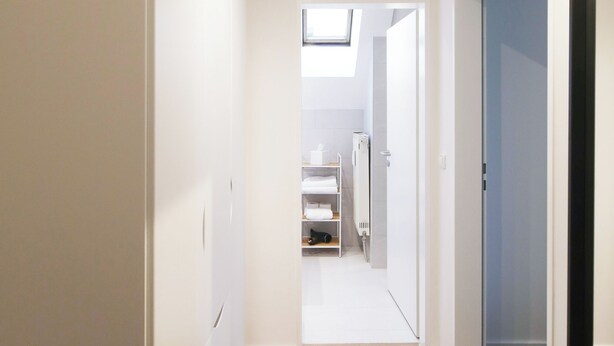 A hallway with wooden flooring, white walls, and a skylight. There is a visible door leading to a bathroom with a towel rack and a toilet.
