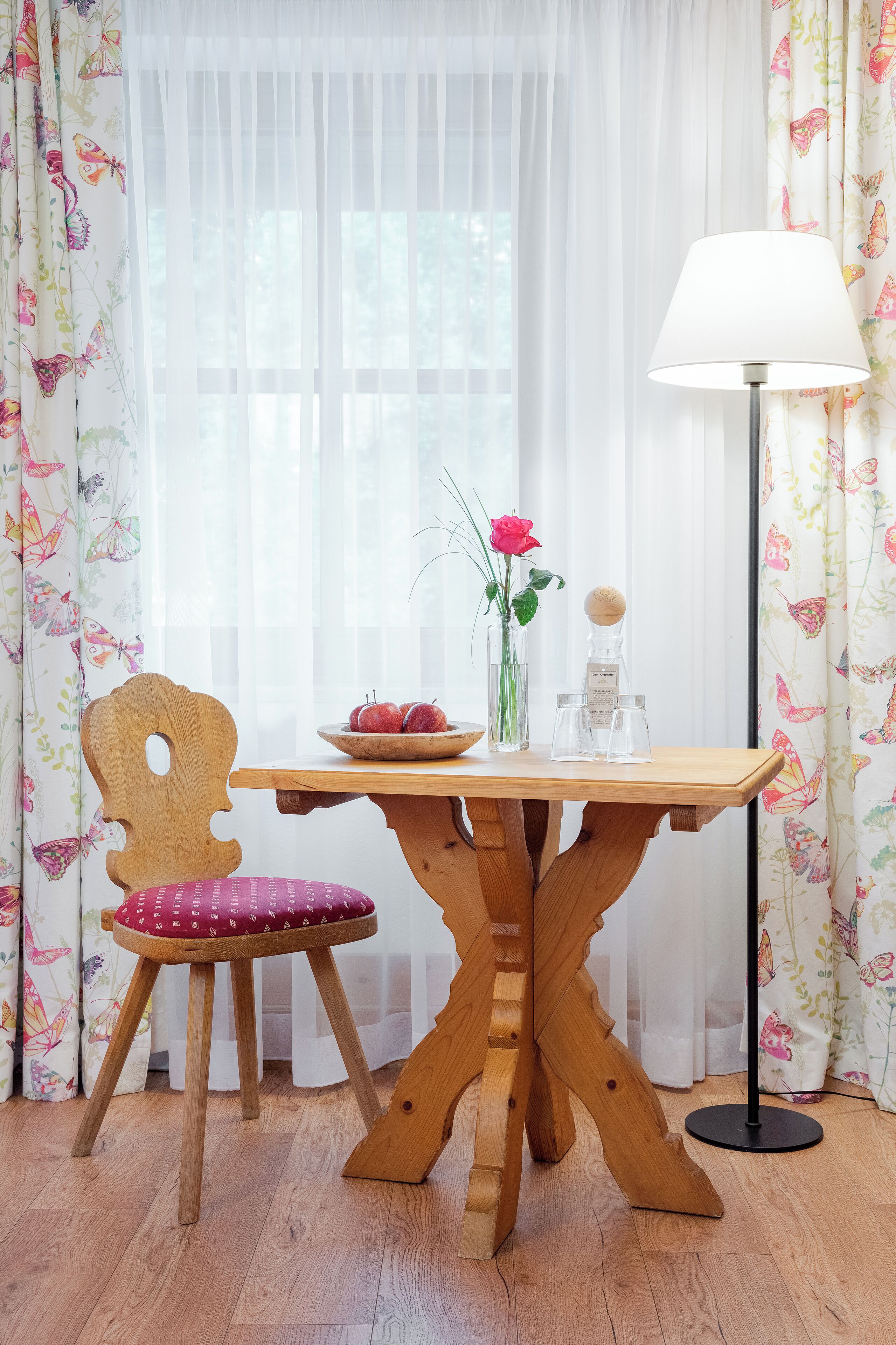 Une table à manger en bois, ornée d’un vase de fleurs et d’un saladier de fruits, une chaise avec un coussin rouge et un lampadaire.