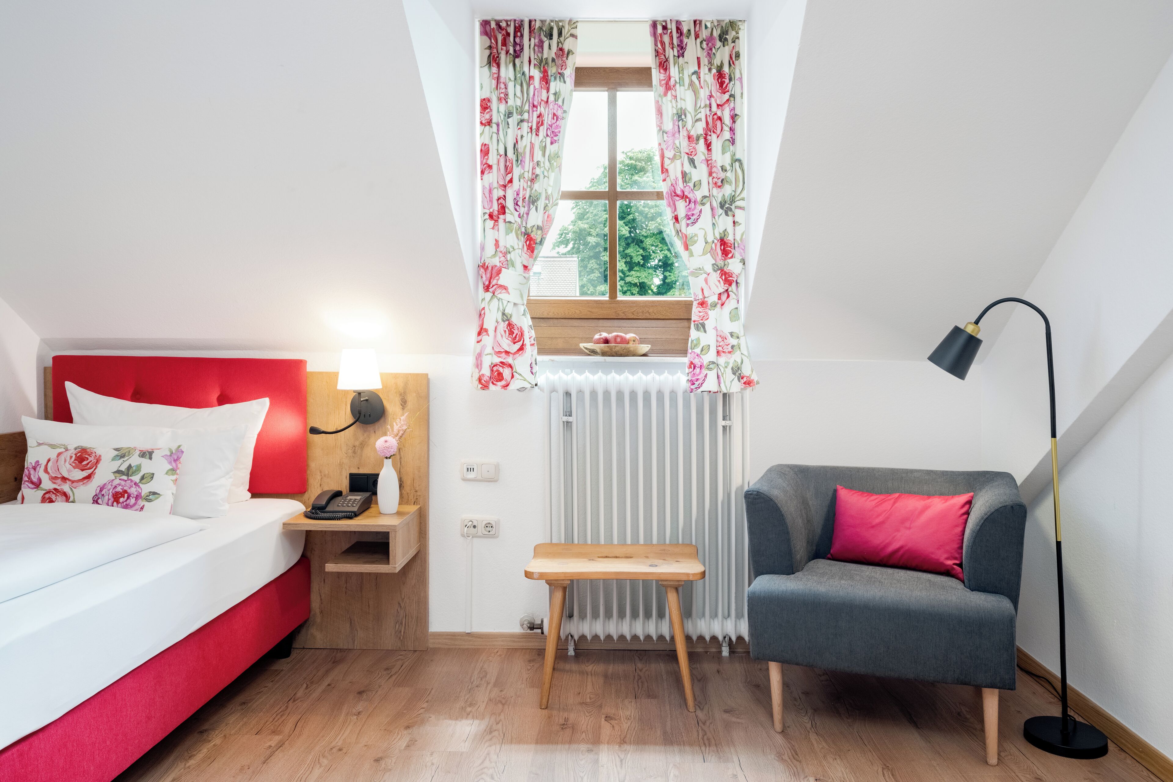 Ein Schlafzimmer mit rotem Kopfteil, einem Nachttisch aus Holz mit Lampe, einem grauen Sofa mit rosafarbenem Kissen, einem kleinen Holzschemel und einem Fenster mit blumigen Vorhängen.