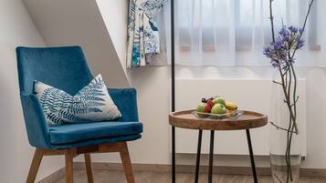 A modern room with a blue chair, a round wooden table with fruits, a floor lamp, and a window with sheer curtains.