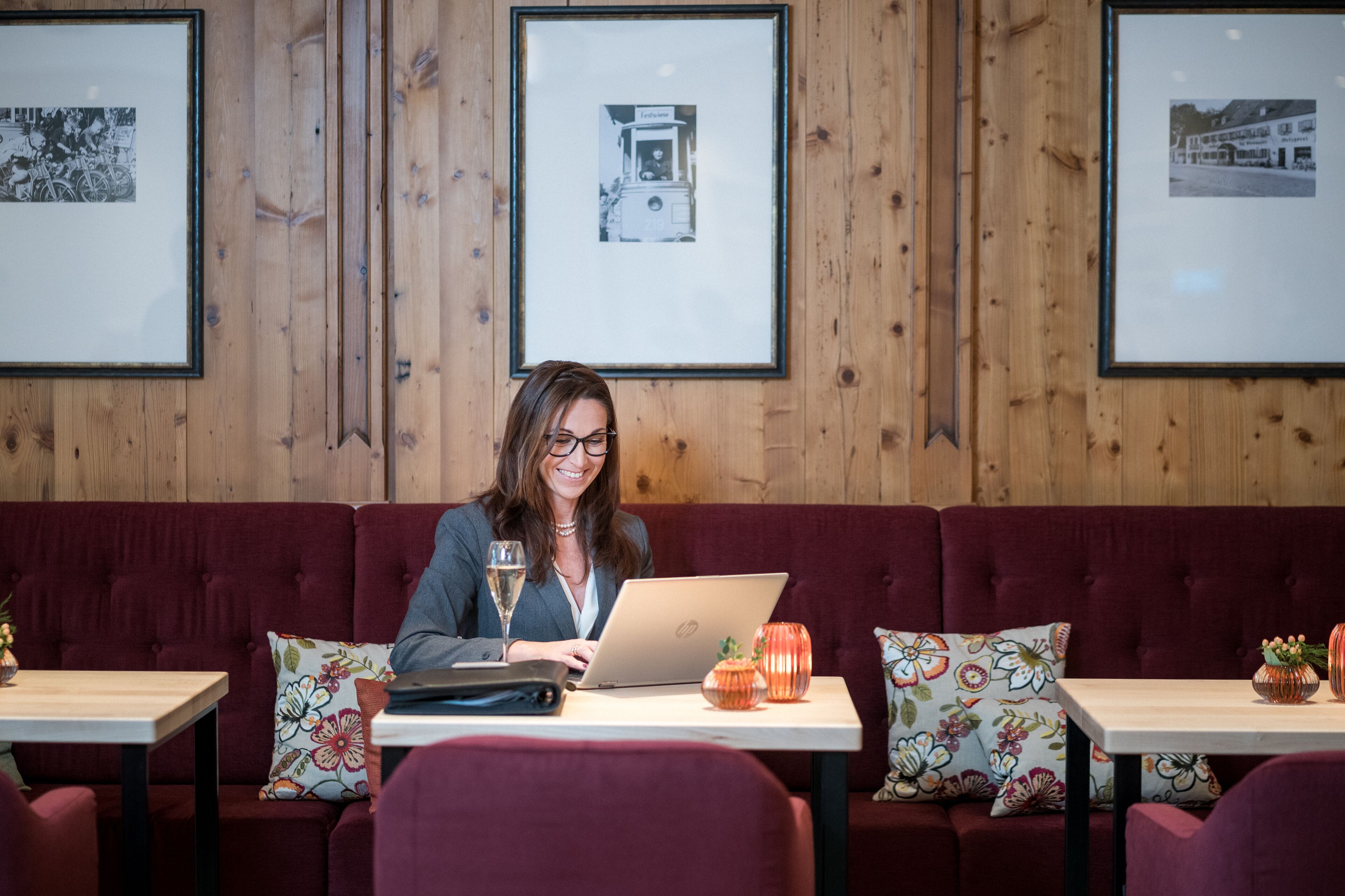 A person seated at a table with a laptop, a glass of water, and decorative items on a wooden wall with framed pictures.