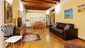Un salon avec un canapé, une table à manger entourée de chaises, une cuisine à l’arrière-plan, des poutres en bois au plafond et des œuvres d’art accrochées aux murs.