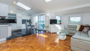 Een moderne woonkamer met een houten vloer, een glazen salontafel, een bank en een televisie. Er is een keuken met witte kasten en een eettafel.