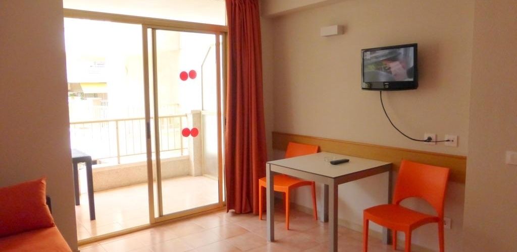 A room with a sliding glass door to a balcony, a table with two chairs, and a television mounted on the wall.