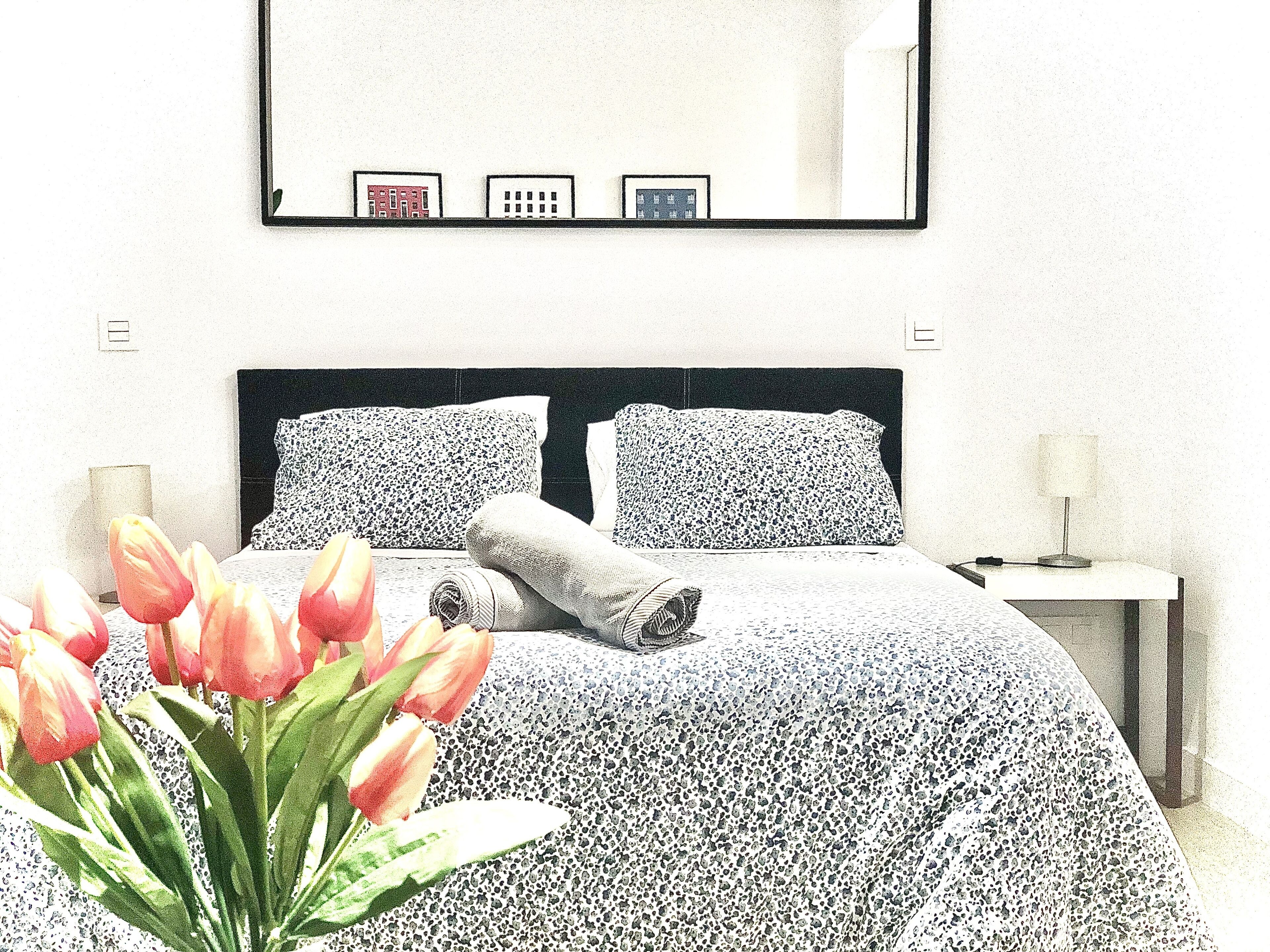 Un dormitorio con cama, almohadas, una mesita de noche, una lámpara y un espejo montado en la pared.