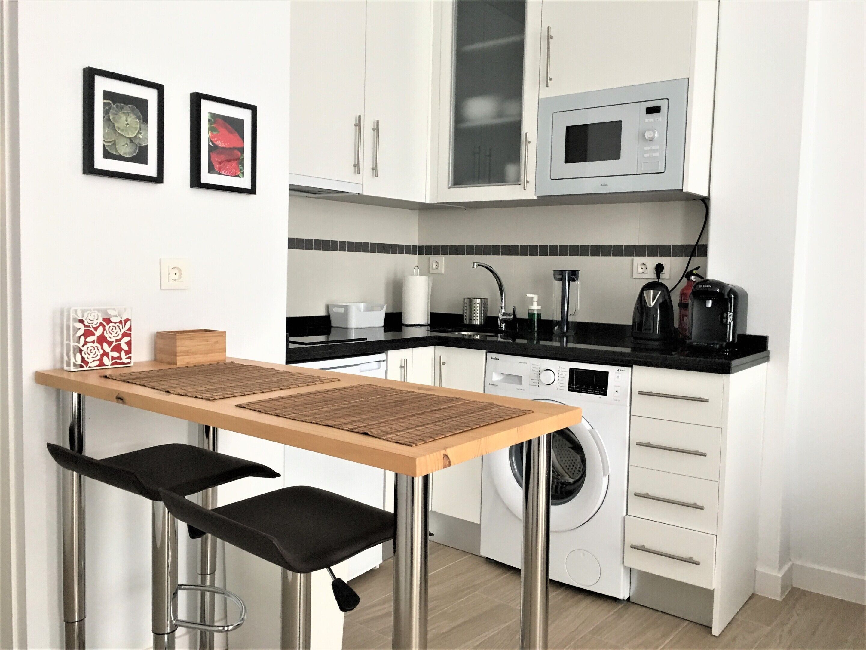 Una cocina compacta con armarios blancos, microondas, lavadora y un pequeño comedor con mesa y sillas.
