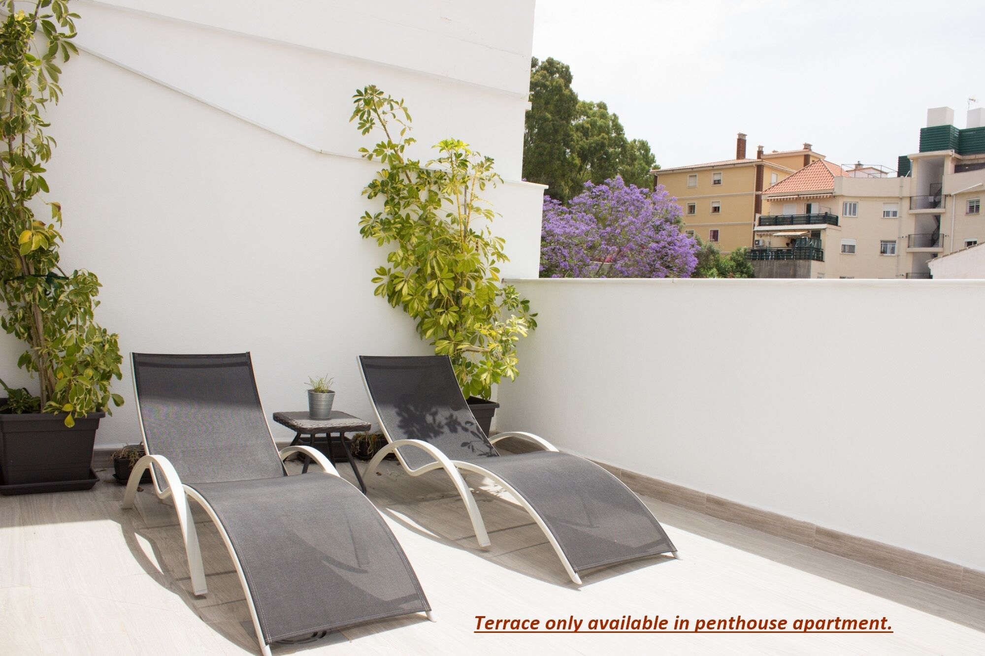A terrace with two lounge chairs and potted plants, surrounded by a white wall and a view of buildings.