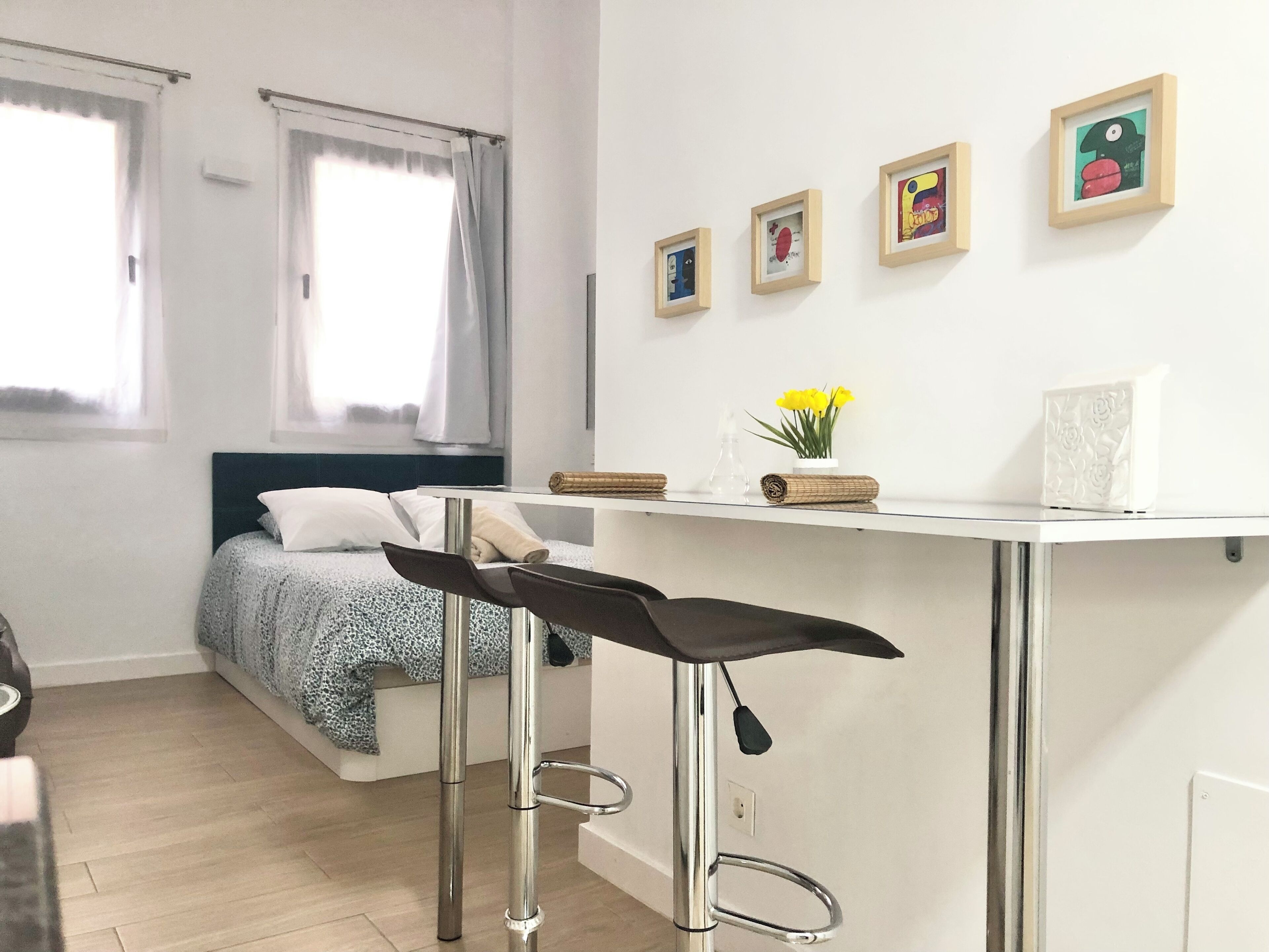 Een kleine slaapkamer met een bed, een klein bureau en een stoel. Er hangen afbeeldingen in lijsten aan de muur en een raam met gordijnen.
