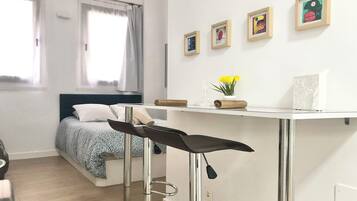 Een kleine slaapkamer met een bed, een klein bureau en een stoel. Er hangen afbeeldingen in lijsten aan de muur en een raam met gordijnen.