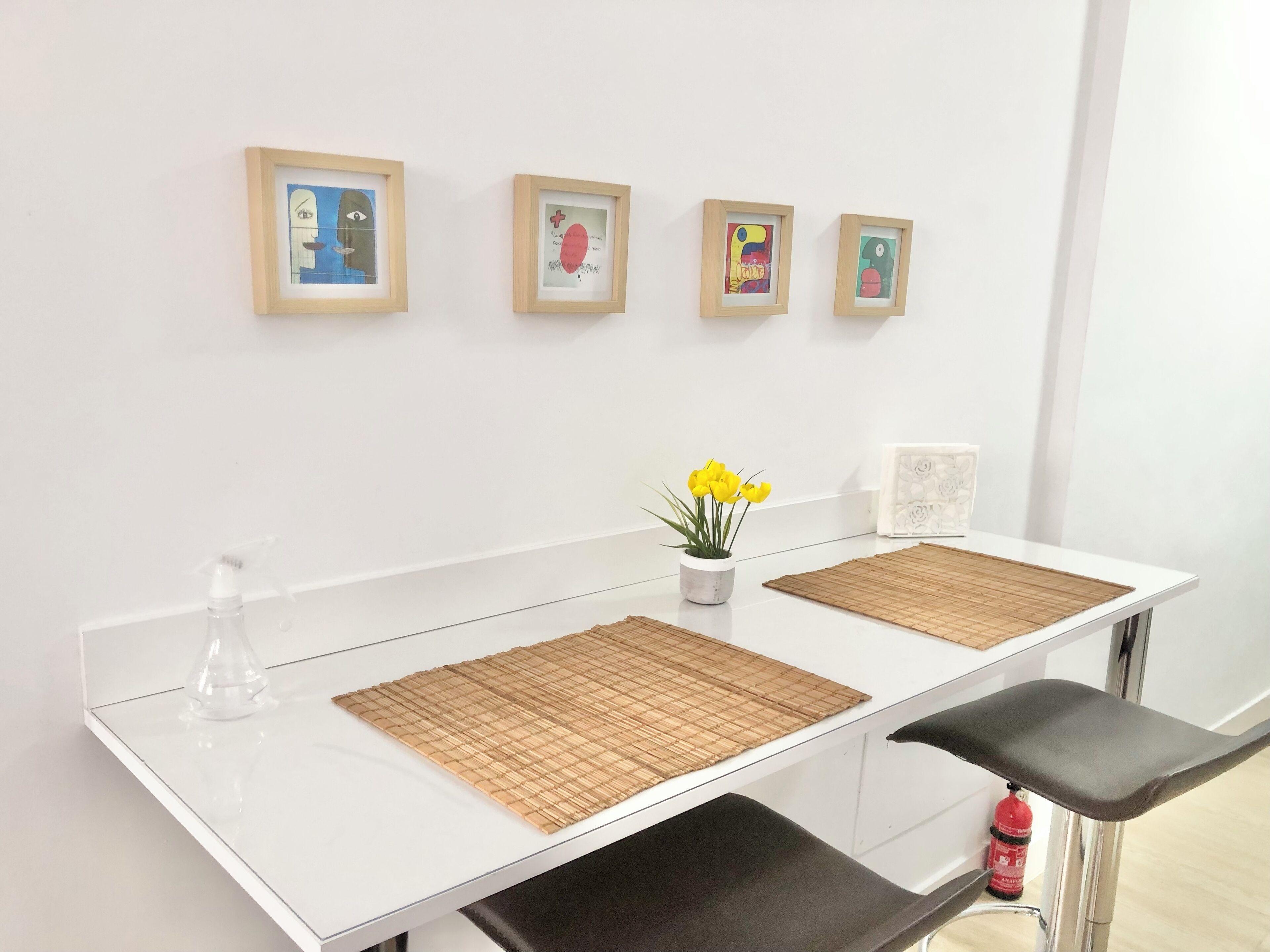 Un mostrador de recepción blanco con una superficie de vidrio transparente, una pequeña planta en maceta con flores amarillas y cuatro cuadros en la pared.