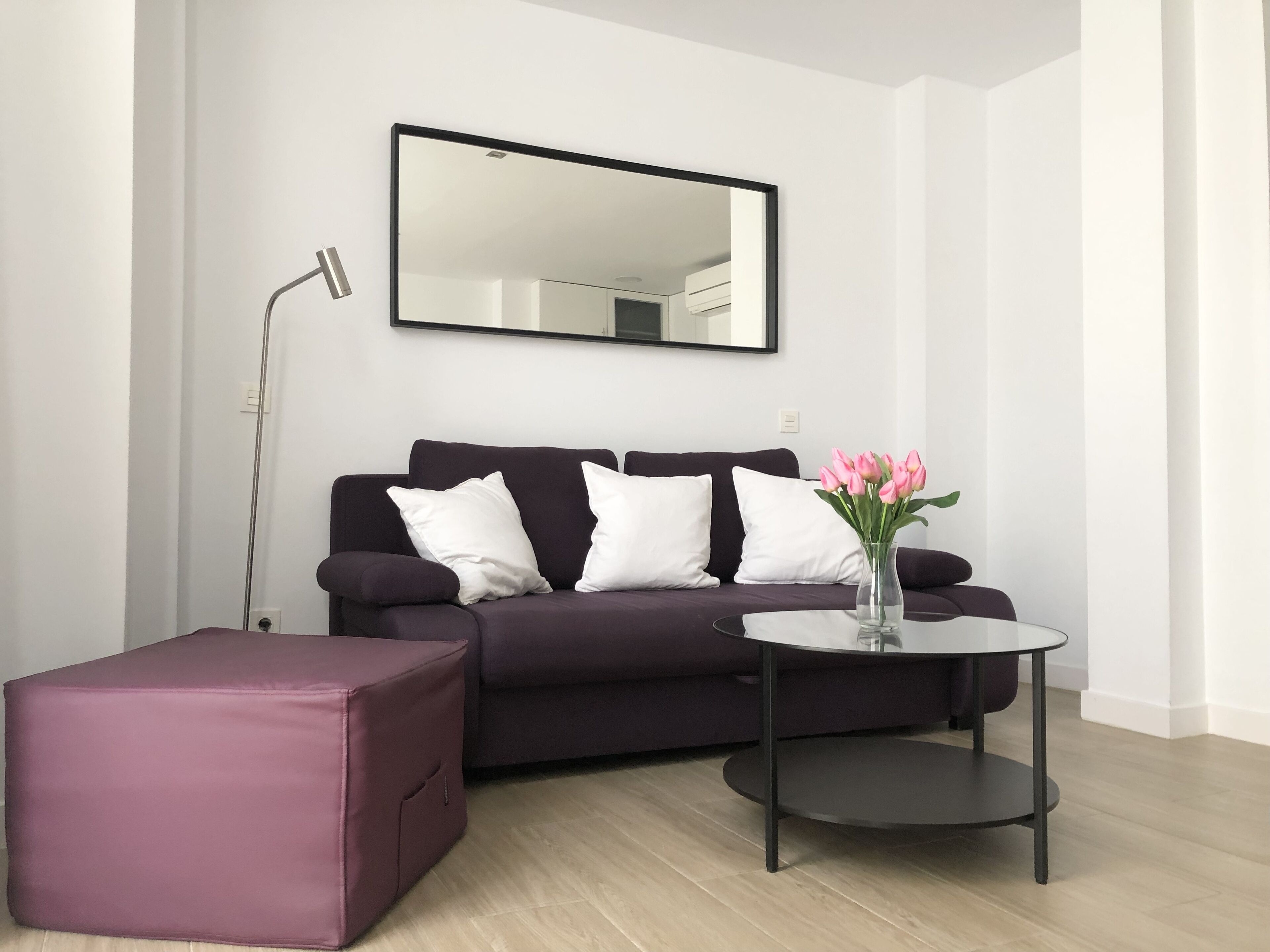 A modern living room with a dark purple sofa, white cushions, a glass vase with pink flowers, and a round black side table.